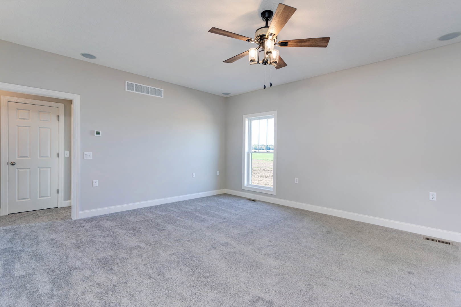 Carpeted room with white walls, ceiling fan with lights, large window overlooking grassy yard, white door with silver handle, wall vent, simple molding.