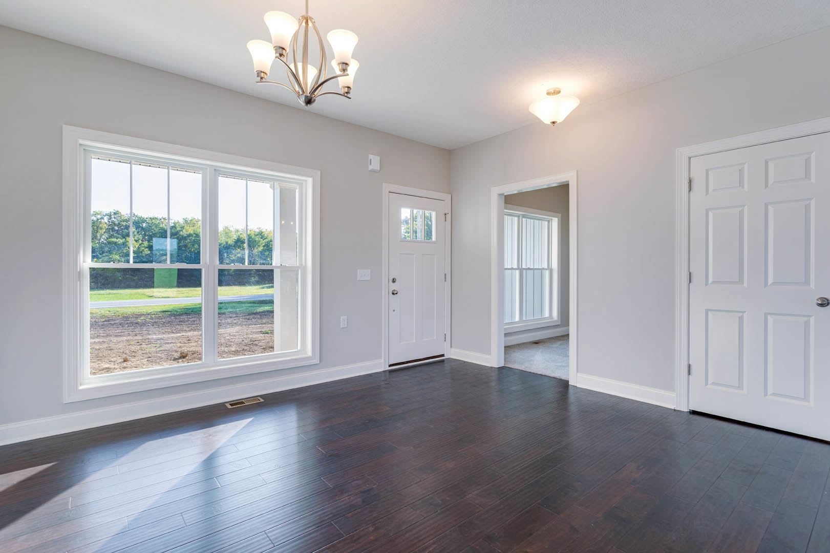 Dark wood flooring and white walls in a room featuring a white door with a silver handle, large windows overlooking a dirt field, and a close-up of a chandelier.