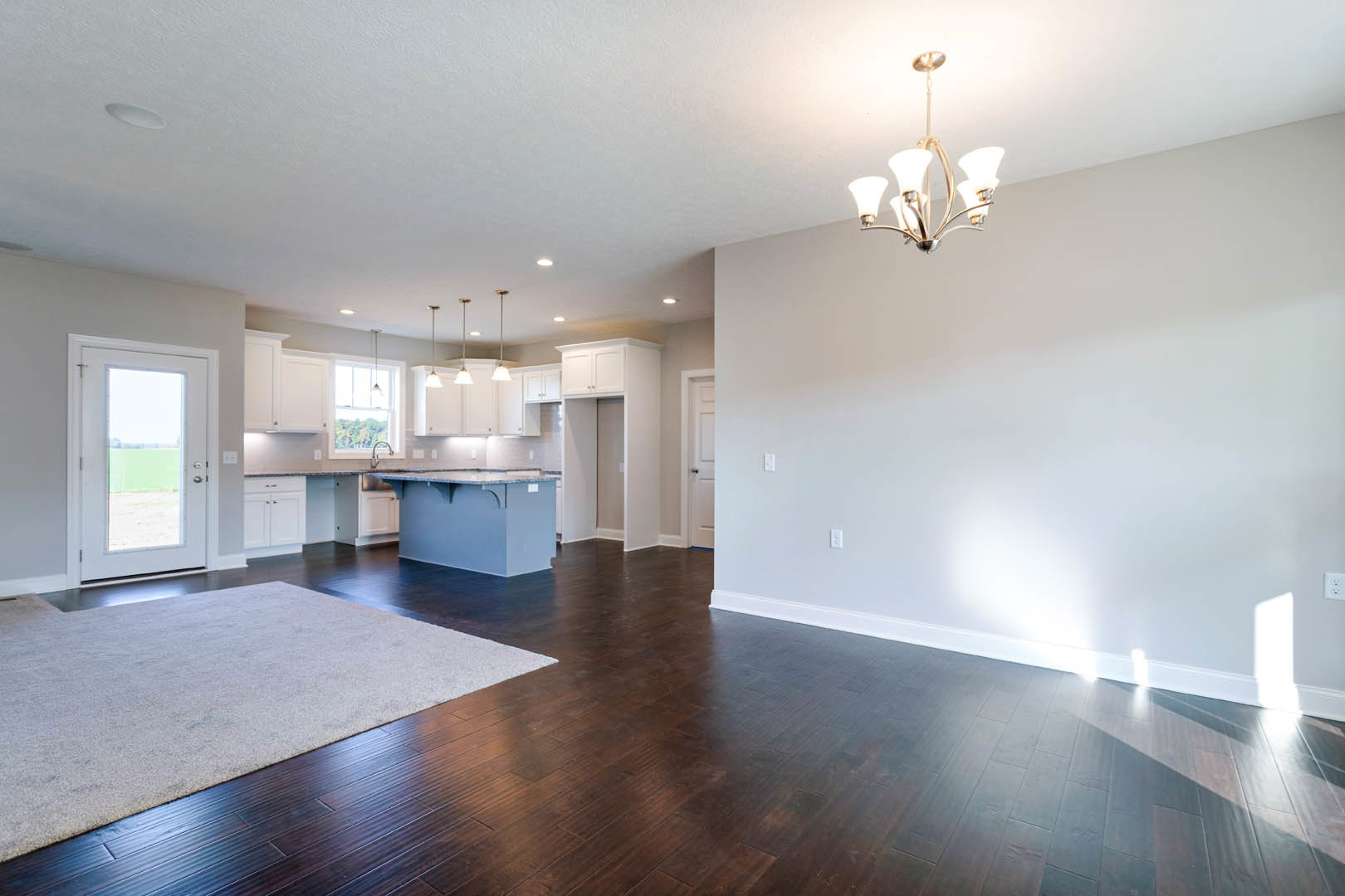 Open-concept kitchen and dining area with dark wood flooring, glass-paneled door, modern chandelier, light-colored rug, and stone countertop