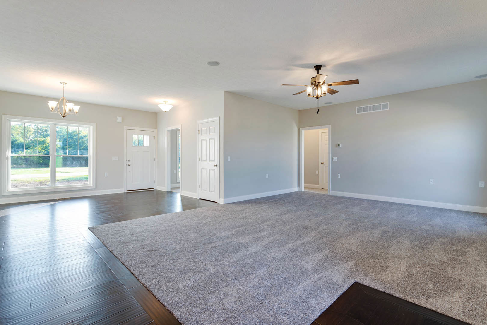 Spacious carpeted room with ceiling fan, white walls, dark wood trim, white door with silver knob, large window overlooking trees