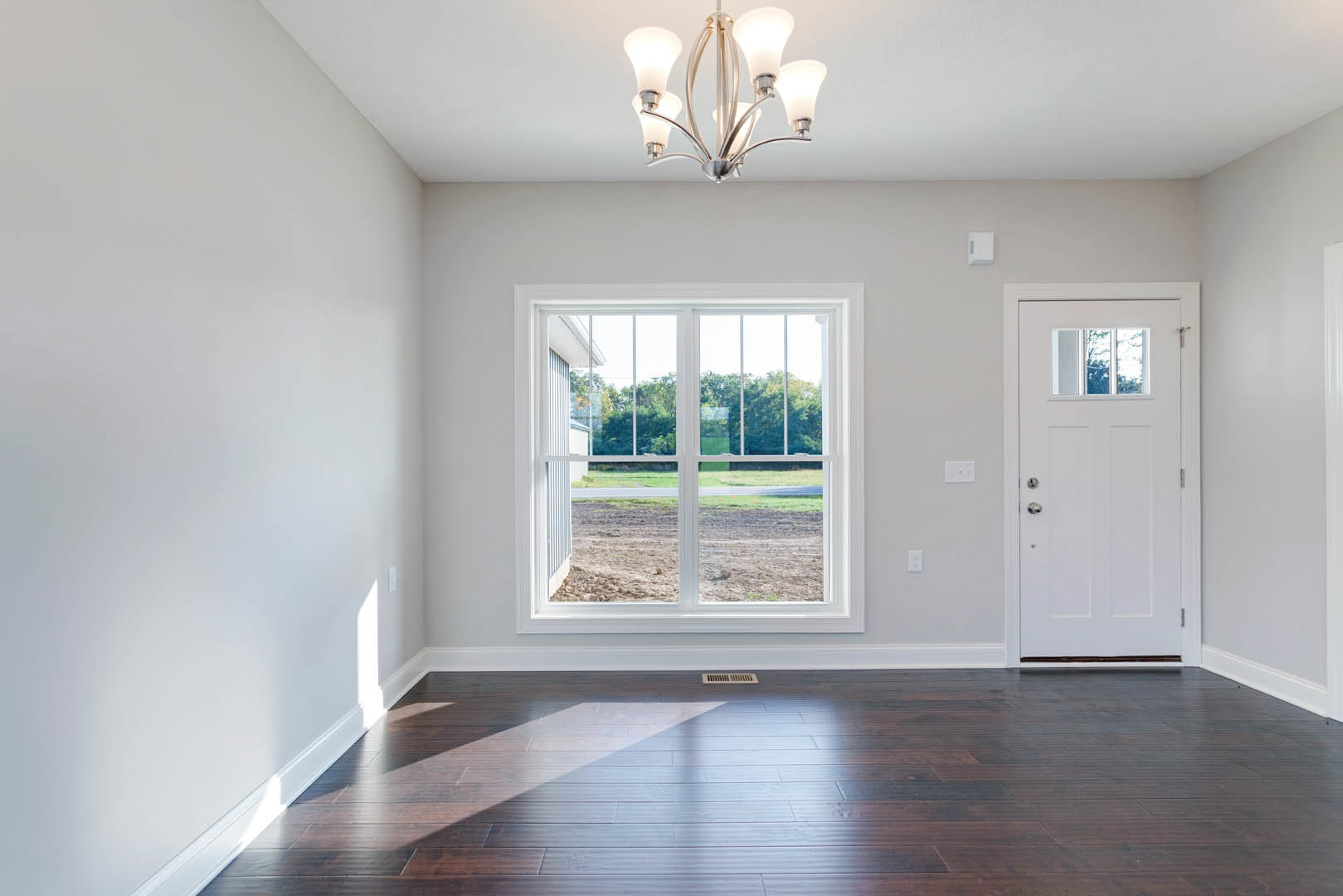 Wood flooring with a vent, white door featuring a window, large window overlooking trees and a dirt field, close-up of a chandelier, white walls with molding