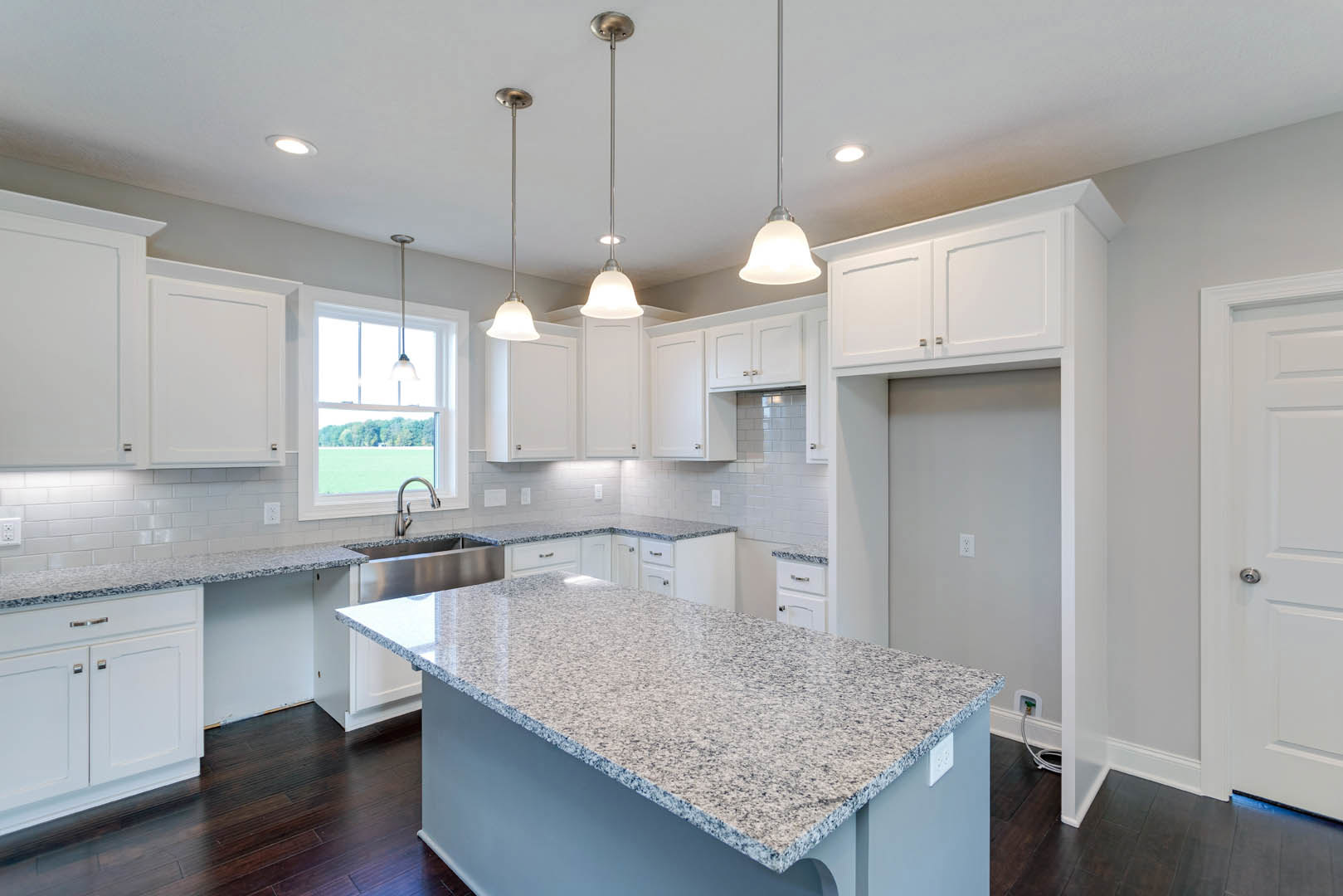 White kitchen cabinets with brushed nickel hardware, granite countertops, stainless steel sink, pendant light fixture, tile backsplash, large window overlooking grassy field.