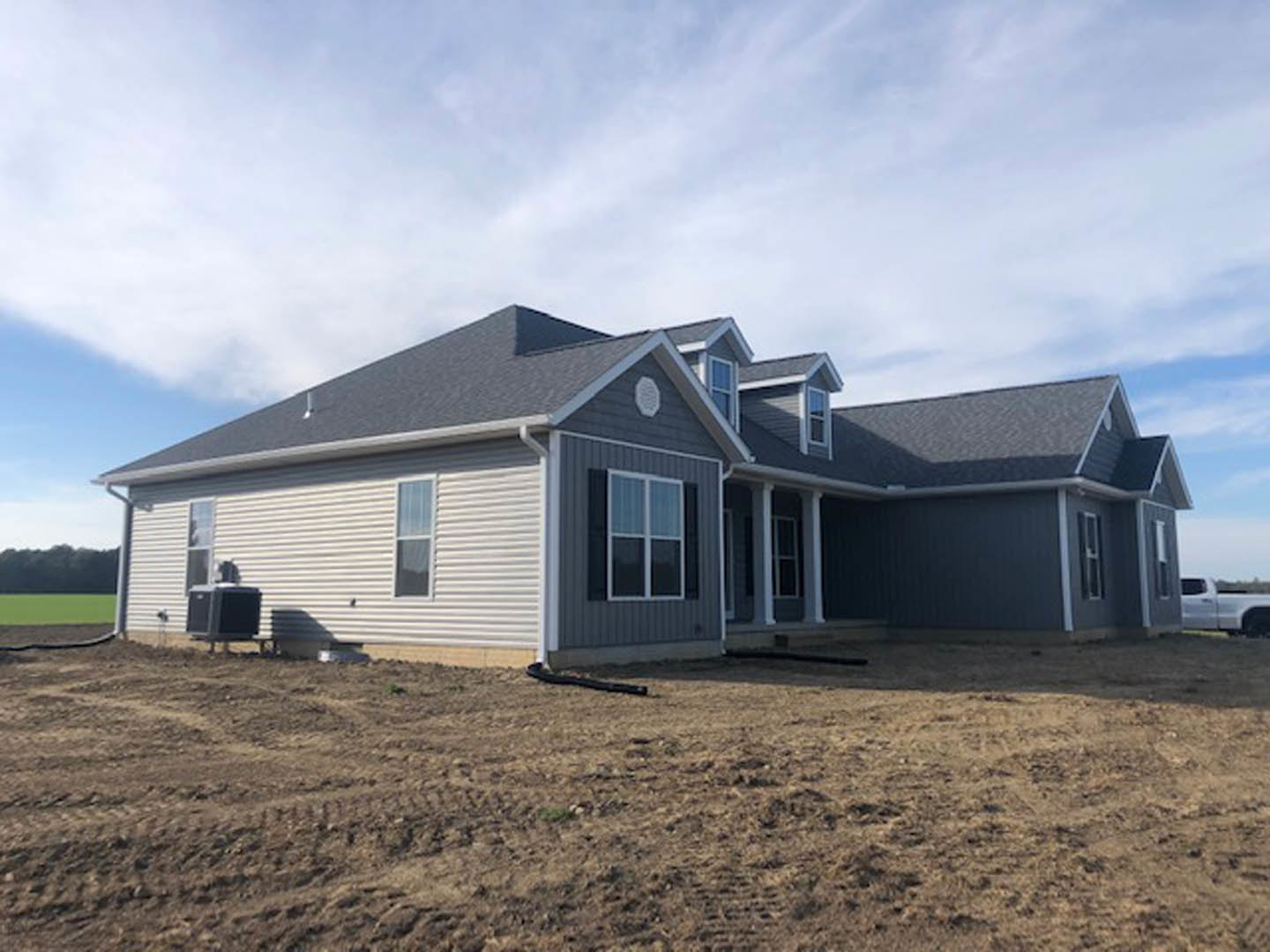 Partially built house with white roof and white-framed windows, surrounded by dirt field and construction materials under clear blue sky; white truck parked nearby, black hose