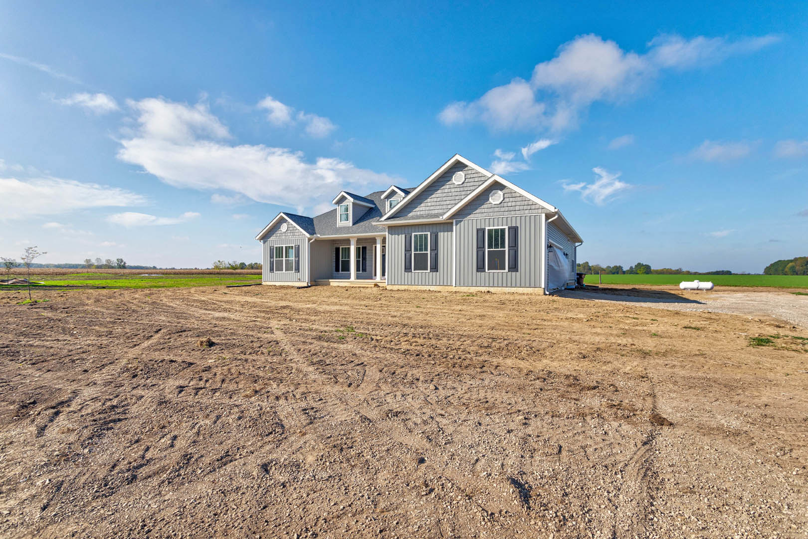 Two-story home with white columns and large windows, set against a blue sky, unfinished dirt yard with tire tracks in front