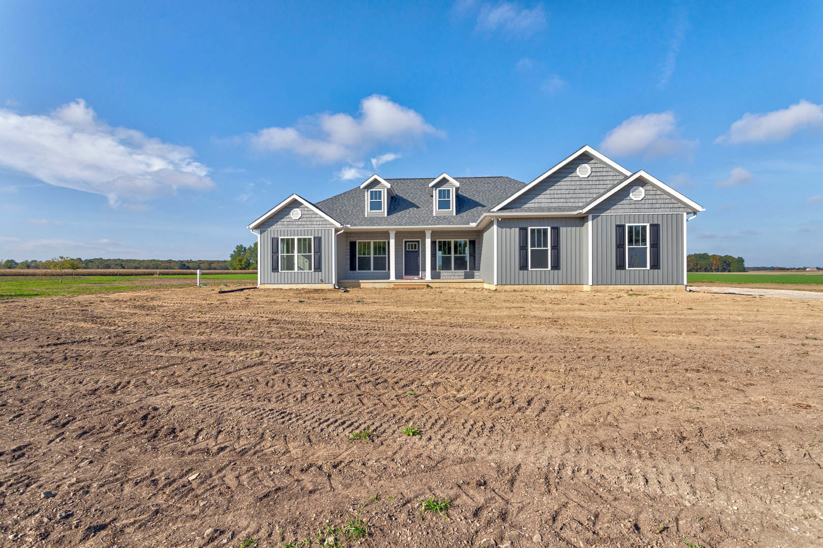 Single-story house with white-trimmed windows, covered porch, and dirt field with tire tracks under a blue sky