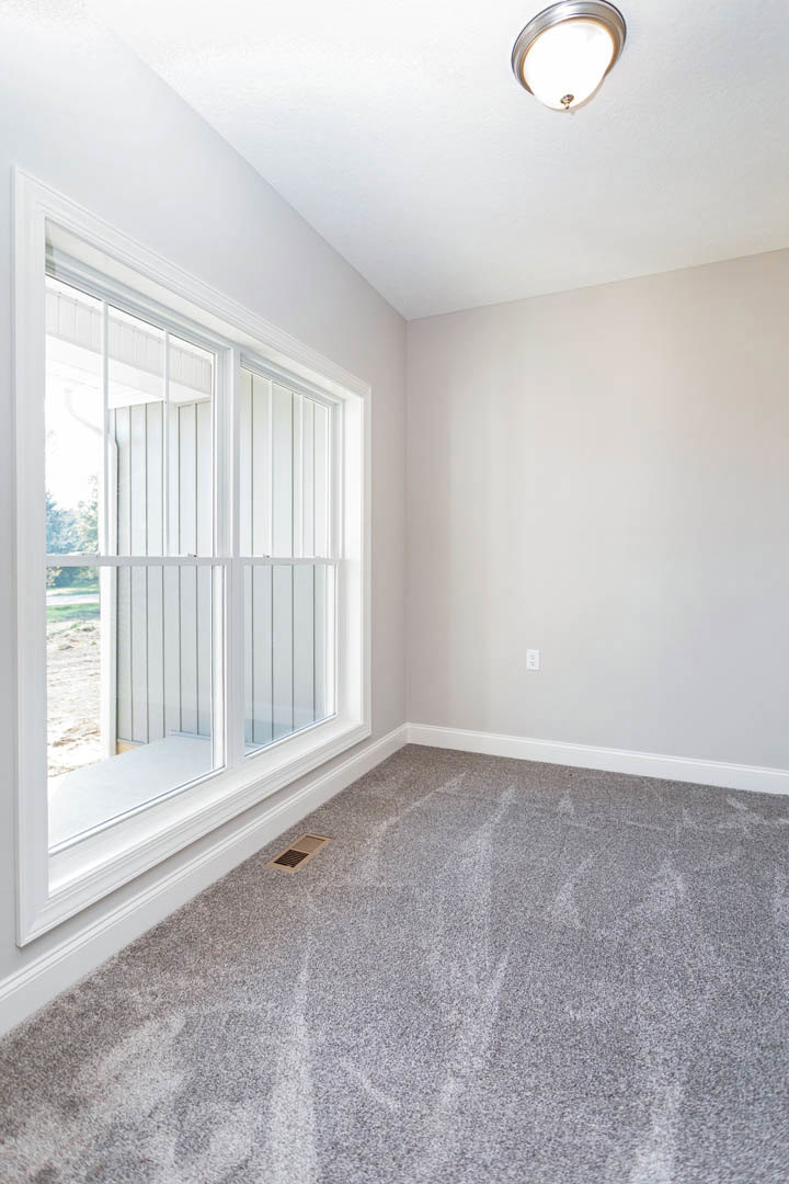 Grey carpeted floor with a corner vent, white walls with electrical outlet, large window with blinds, ceiling-mounted light fixture, simple molding along baseboards