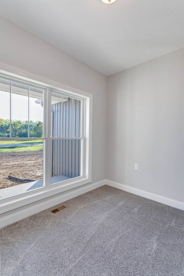 Grey carpeted floor with a corner air vent, large window with white blinds, view of dirt and grass outside, white walls with base molding