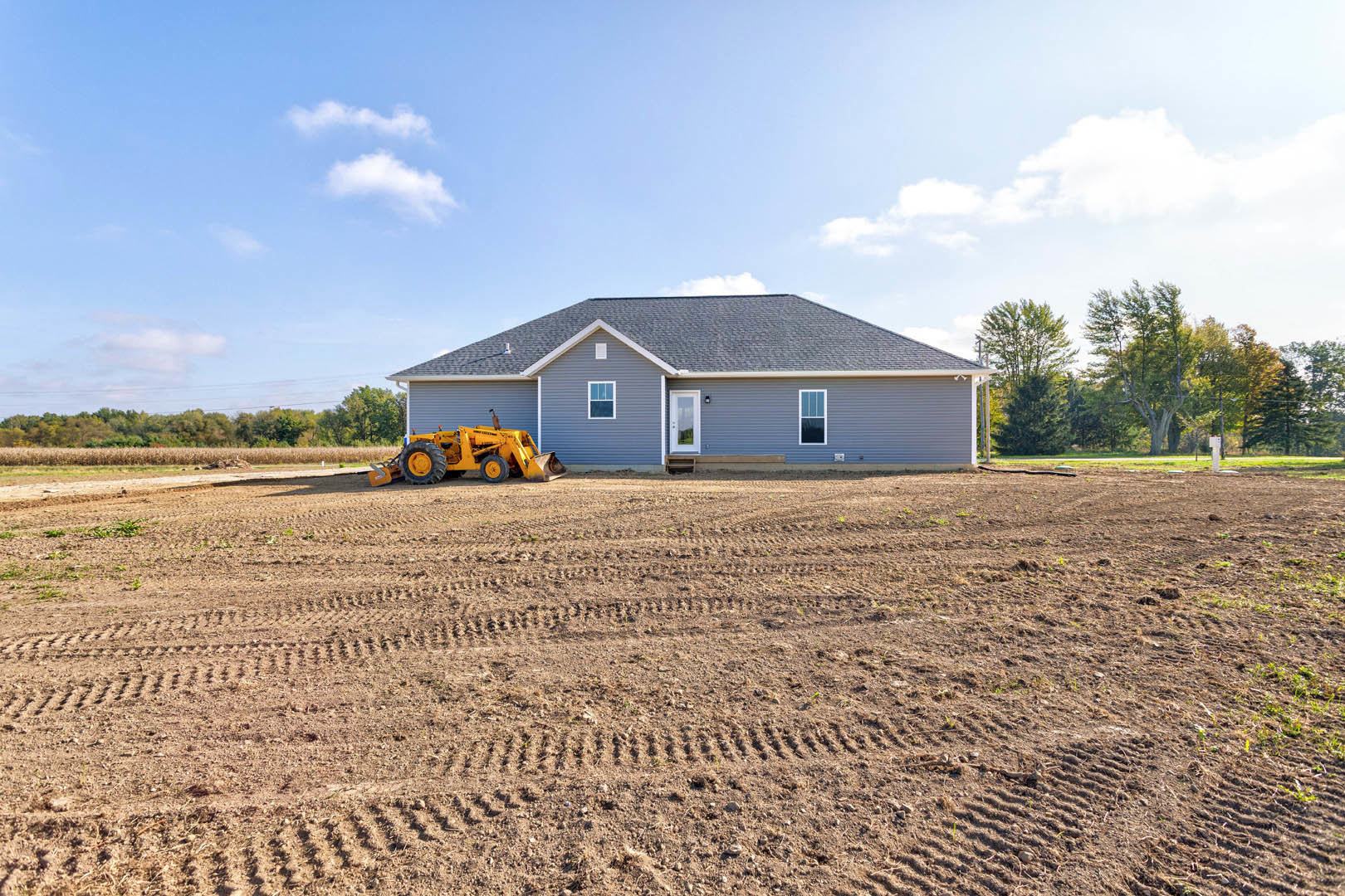 Yellow tractor with large wheels parked on dirt lot in front of single-story house with white door, surrounded by open field and tree under blue sky.