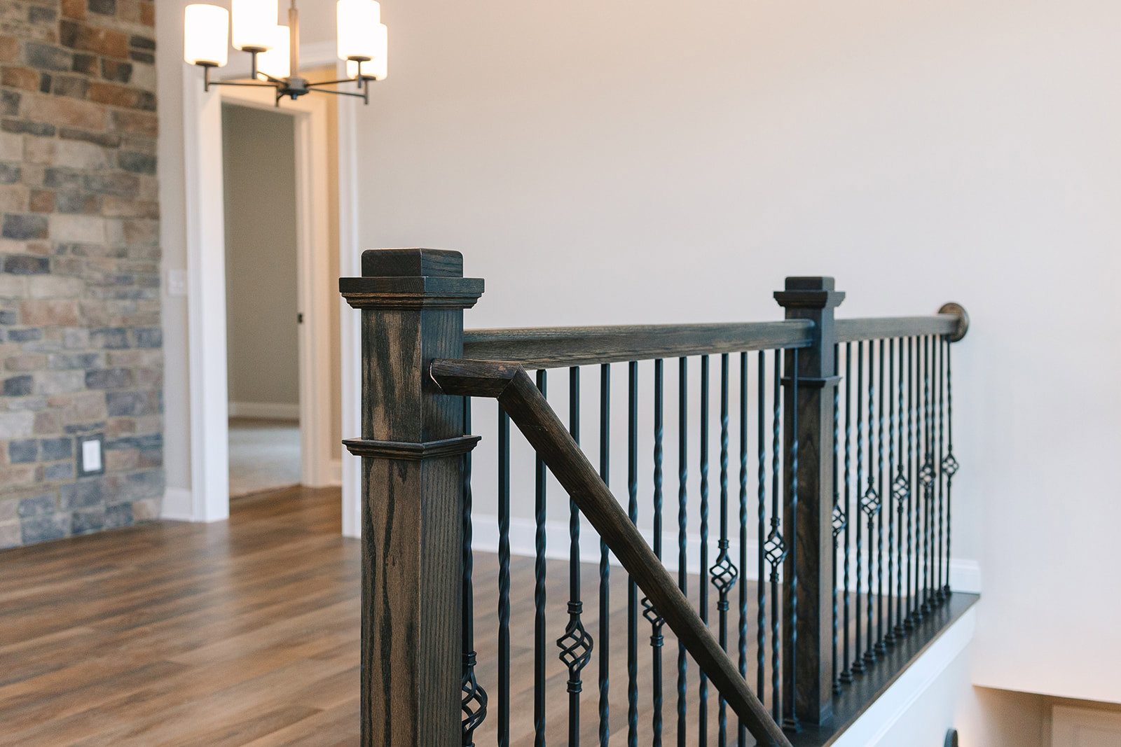 Wooden staircase railing with iron balusters, close-up of wooden post, chandelier overhead, blurred brick wall in background, hardwood flooring