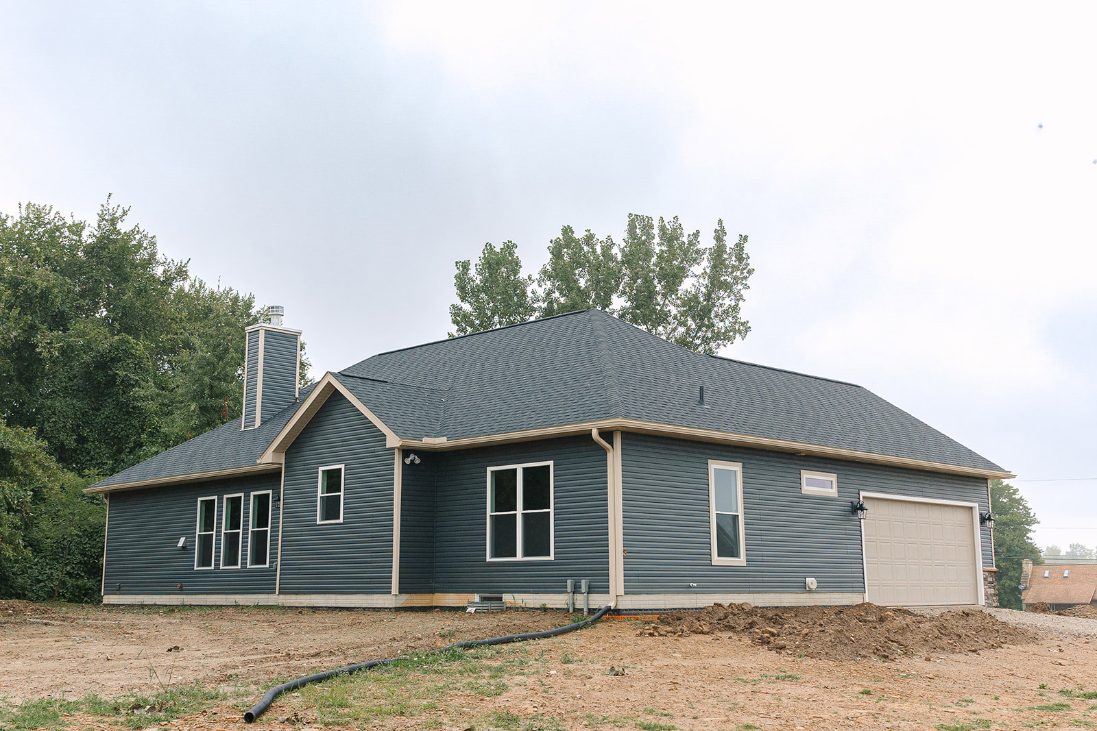 Two-story house with white-framed windows, attached garage featuring a light post, garden hose lying on dirt near the driveway, surrounded by trees and open sky.
