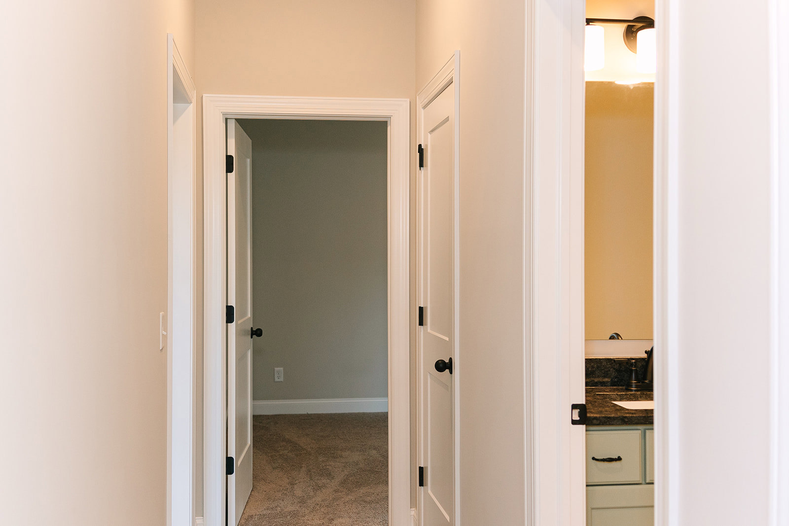 Hallway with white paneled doors, carpeted walls, beige carpet flooring, open door leading to a room, gold-bordered white wall accent, two-lamp ceiling light fixture, close-up of a