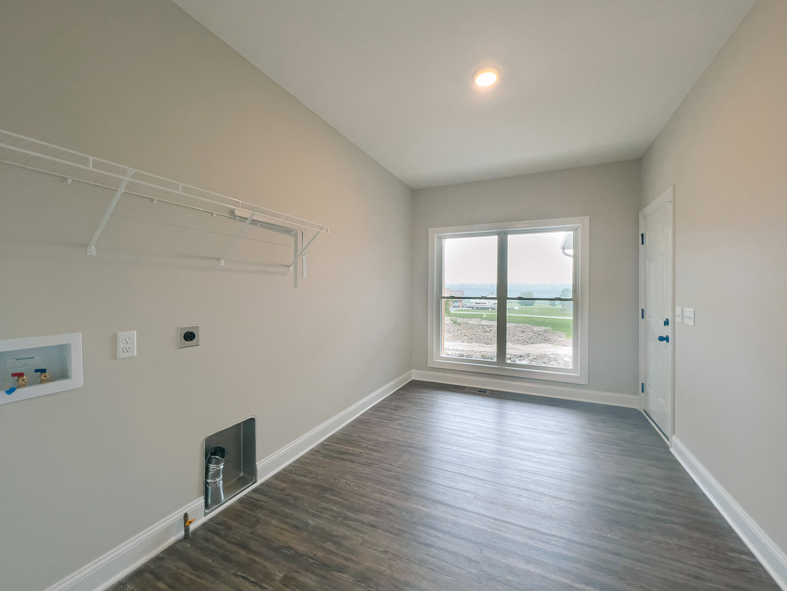 Sunlit room featuring wide wood flooring, white plaster walls, ceiling light fixture, large window overlooking a field, and a white utility box with brass faucets and exposed metal