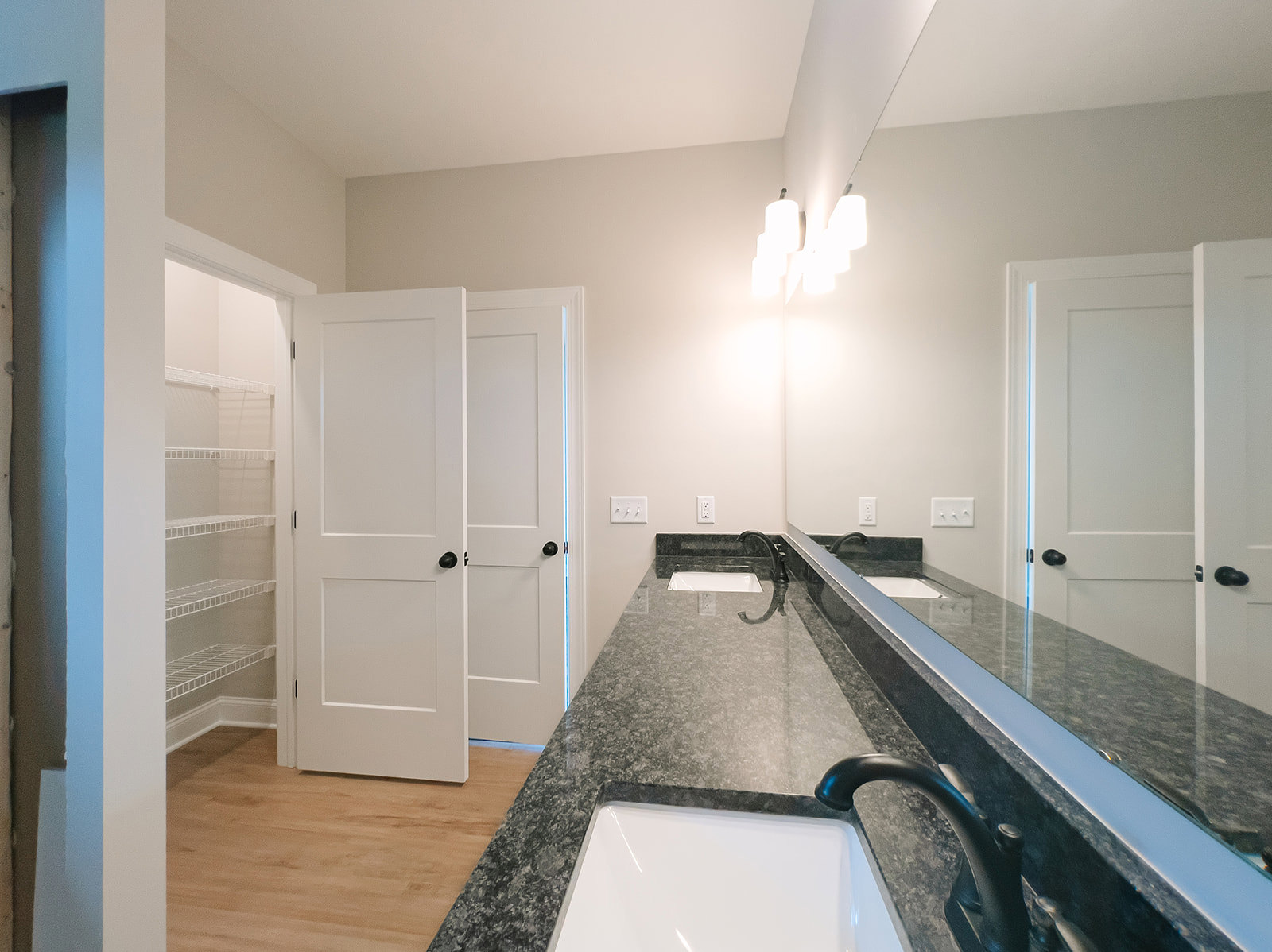 Bathroom with double sinks, wide frameless mirror, white quartz countertop, black hardware, wall-mounted light fixture, white cabinetry, and neutral tile flooring
