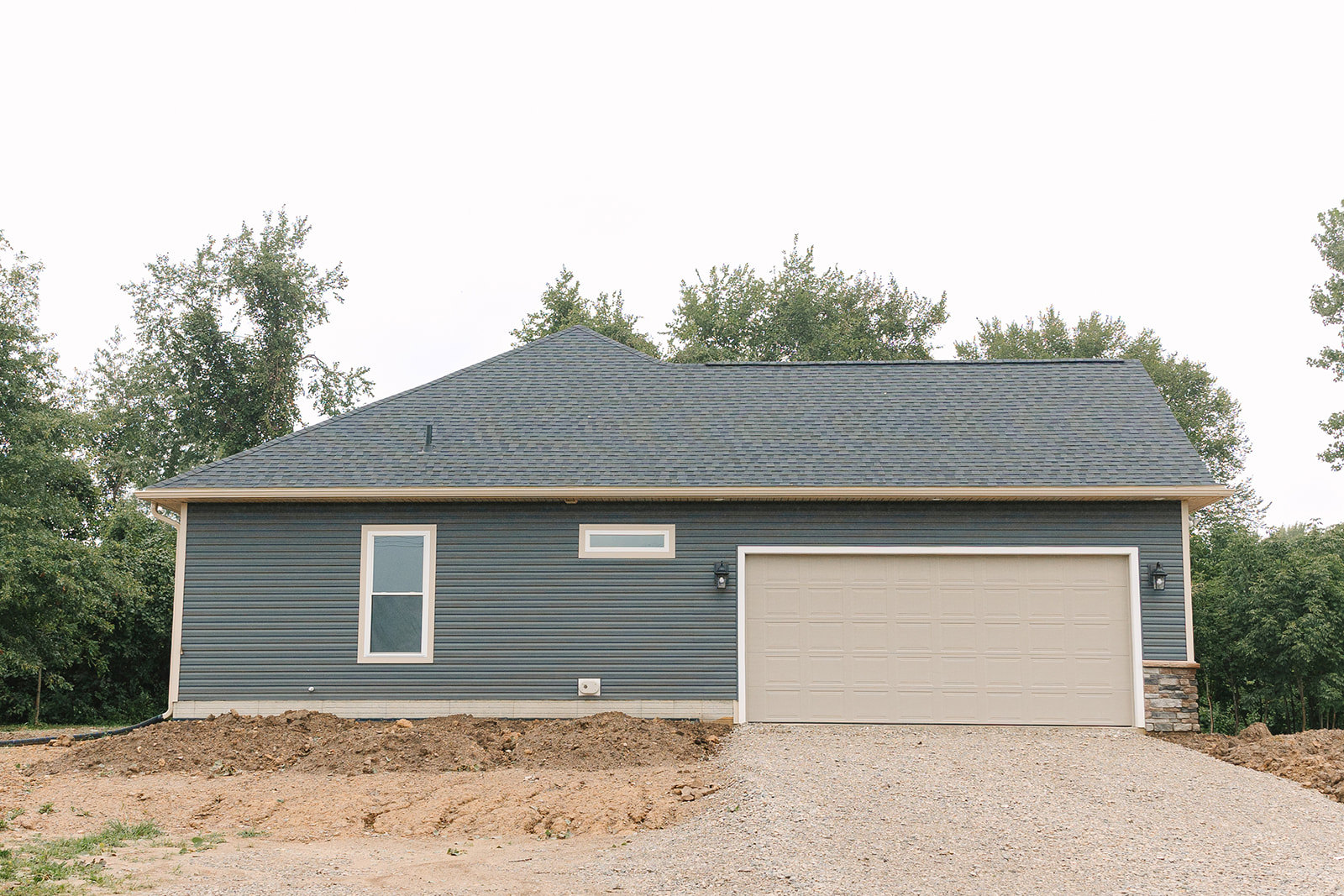 Two-story home with white siding, black roof, attached white garage door, large front window with white frame, pile of dirt and gravel in driveway, surrounded by trees.