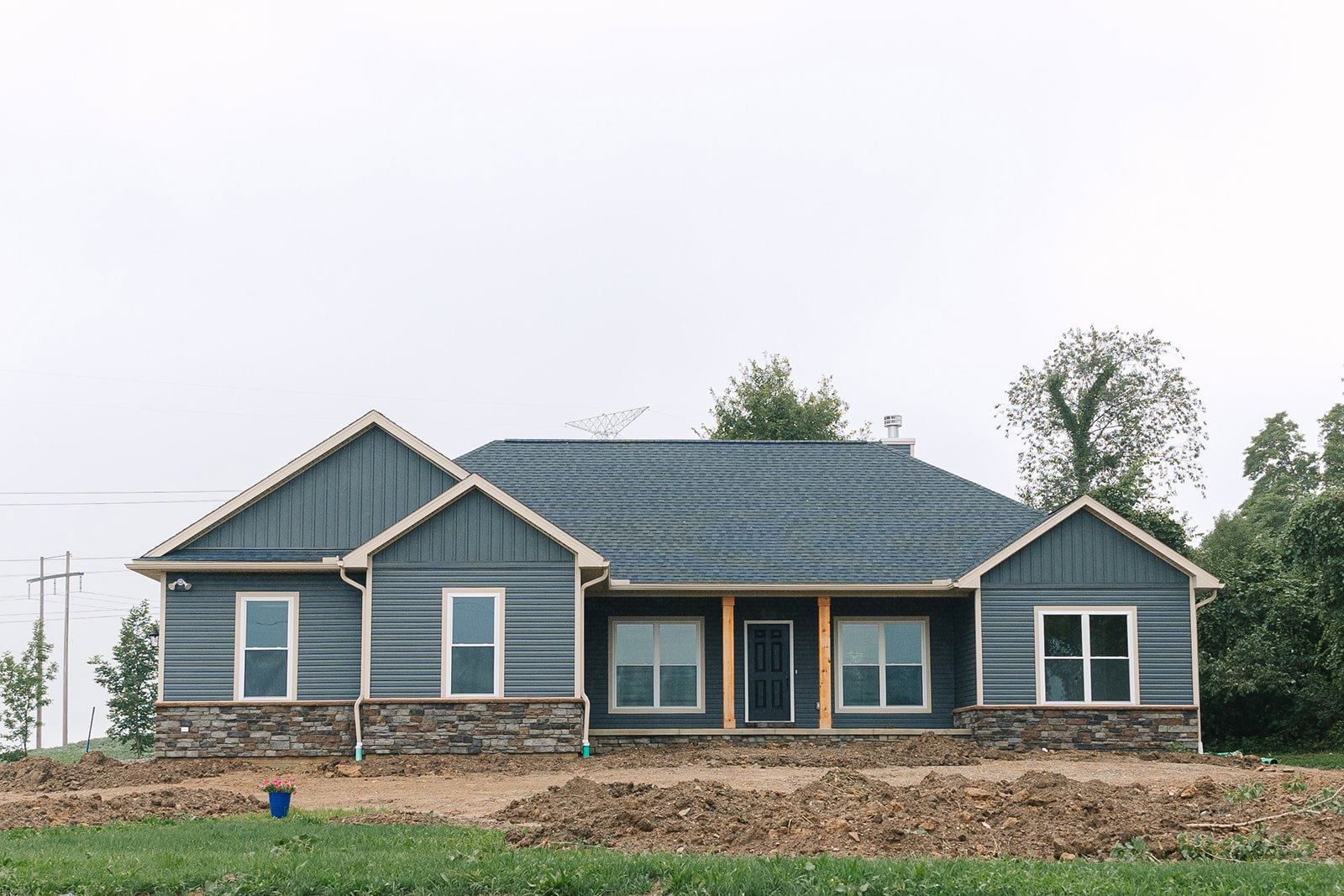 Blue metal roof, brick and stone exterior walls, white-framed windows, black front door with white trim, broken glass in one window, landscaped lawn with trees and grass under a