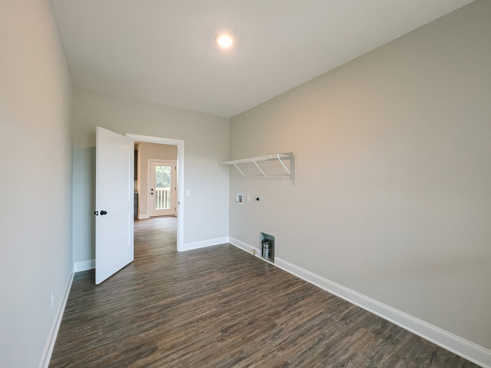 Wood flooring in a room with white plaster walls, ceiling light fixture, open white door featuring glass window and black knobs, silver pipe and white metal bar mounted on wall.