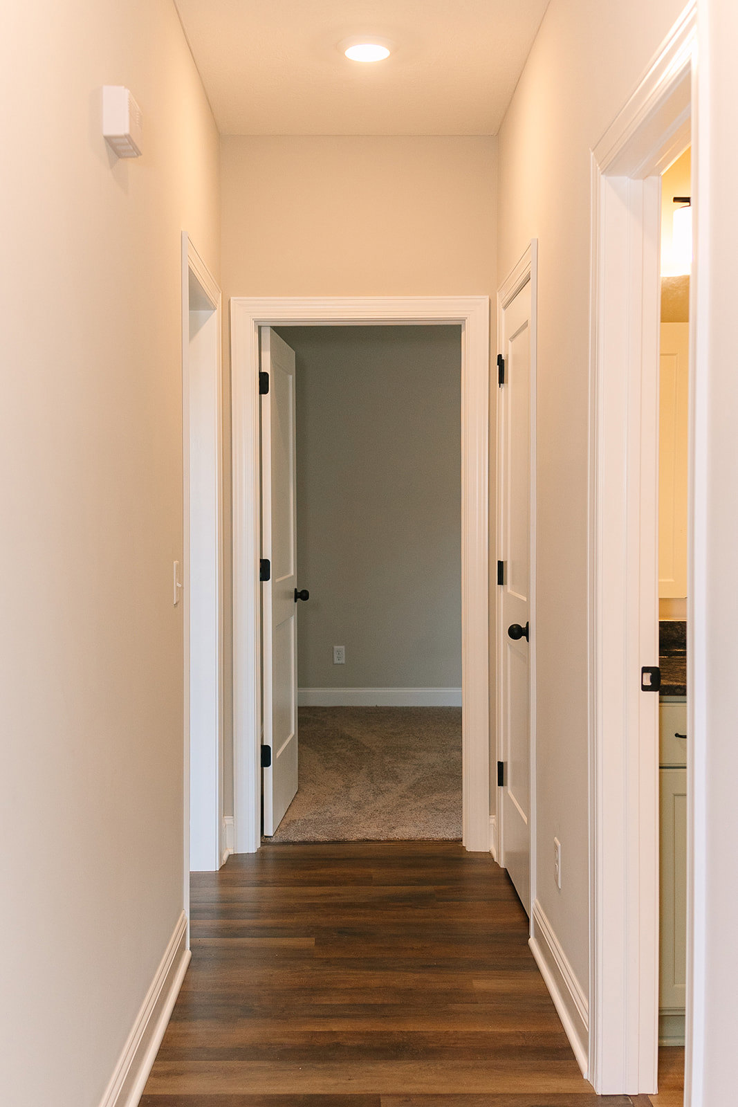 Hallway with white walls, black doors, wood flooring with white trim, ceiling light, white rectangular vent, open door leading to adjacent room