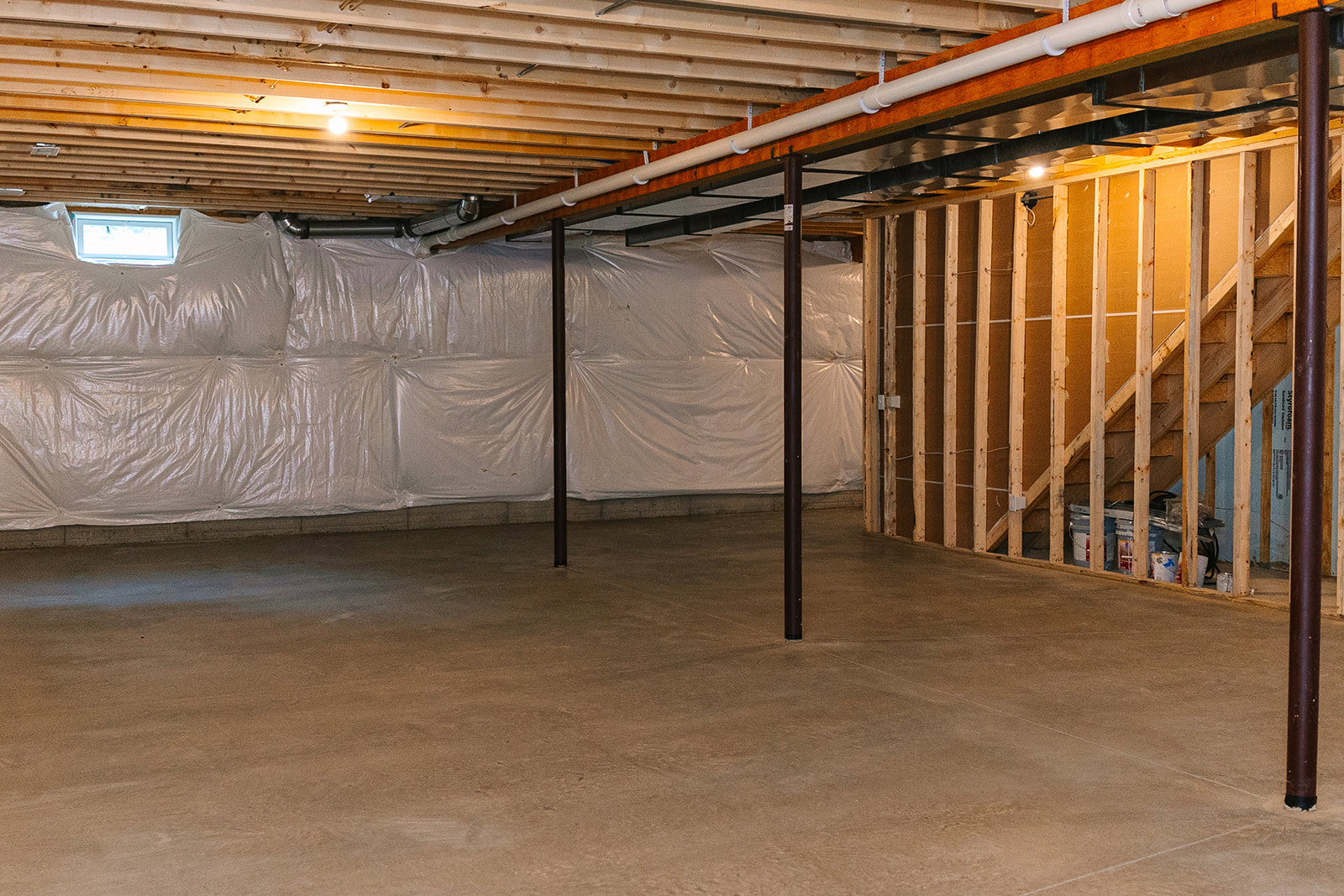 Basement with white plastic sheeting covering ceiling and walls, exposed wooden beams, white pipe along wall, metal support pole on concrete floor, close-up window visible.