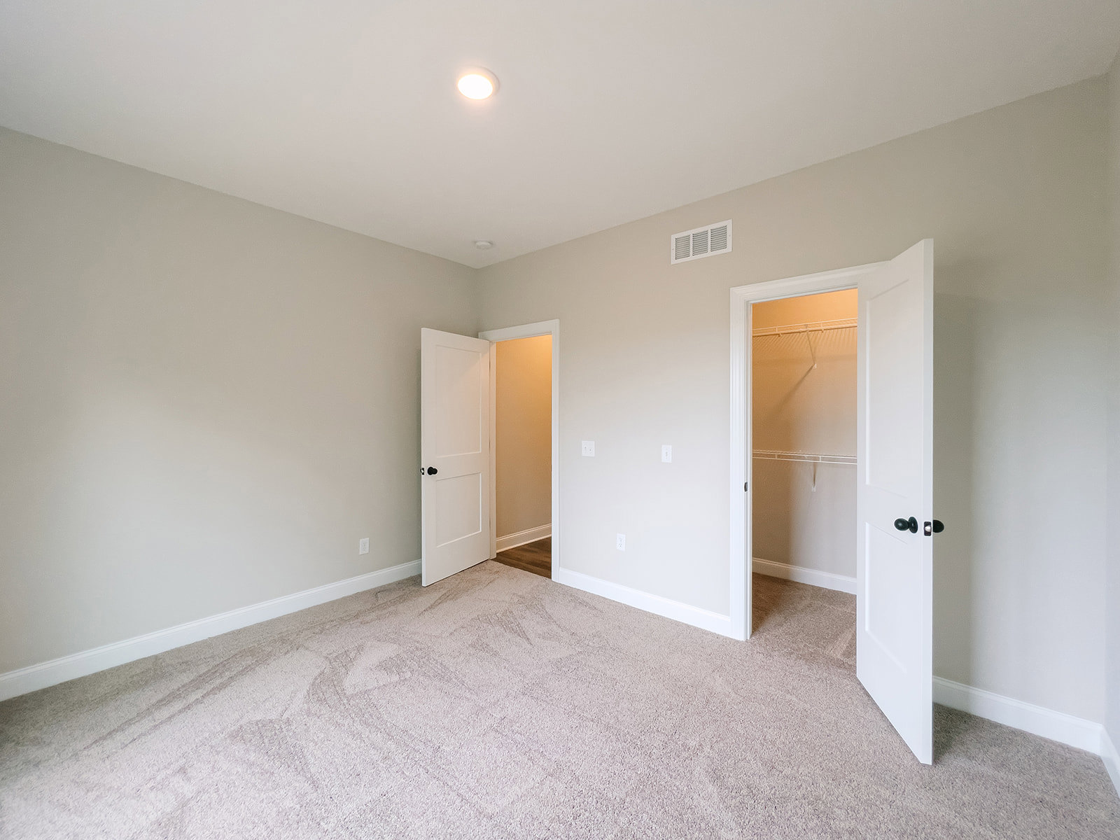 White room with laminate flooring, two open white doors with black knobs, ceiling light fixture, wall vent, and white door frames.