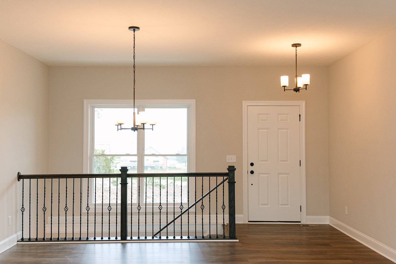 White paneled door with black hardware beside black metal railing, warm wood laminate flooring, modern chandelier hanging near window, neutral walls
