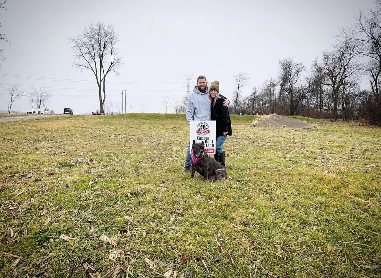 Couple standing beside a sign in a grassy field with a dog sitting nearby, surrounded by trees and open meadow under a clear sky