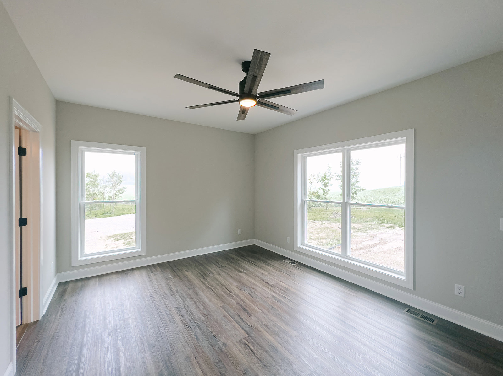 Ceiling fan with light fixture, wood flooring, large windows showing grass and trees, white walls, close-up of brushed metal door handle