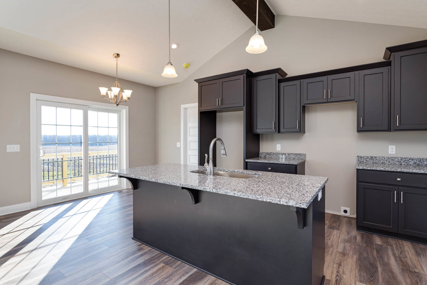Granite kitchen island with built-in sink, wood flooring, white cabinetry, stainless steel appliances, sliding glass door overlooking open field, modern chandelier hanging from