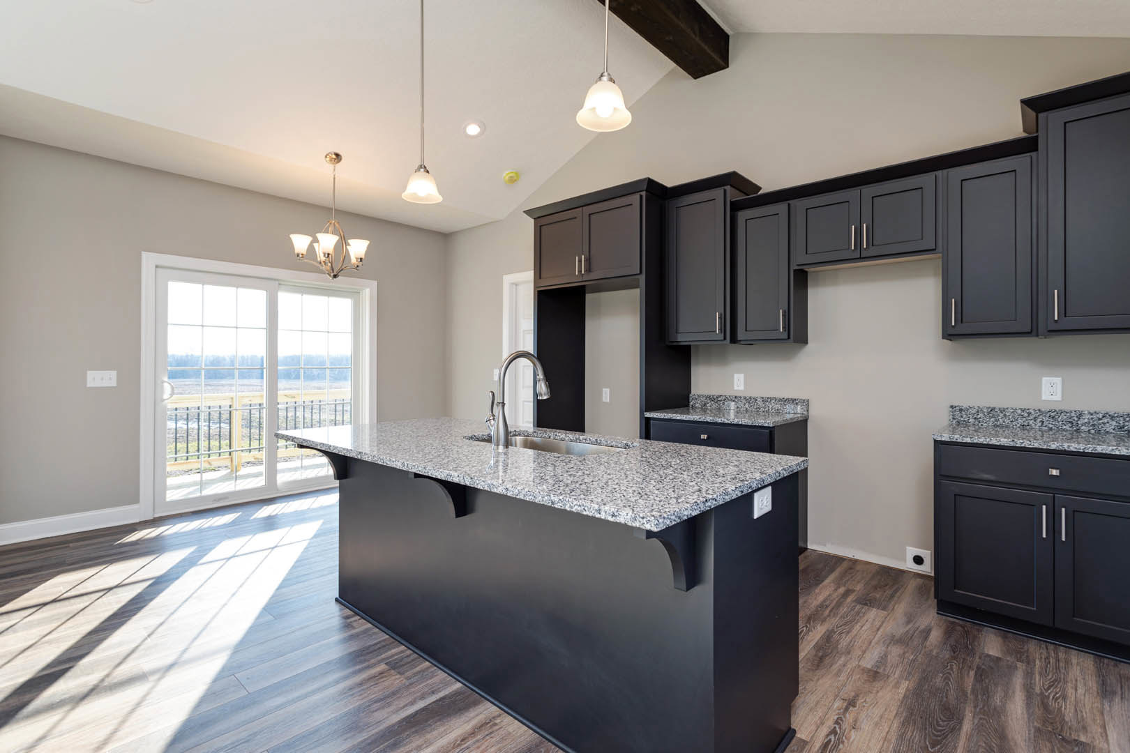 Spacious kitchen featuring a large central island with stone countertop, wide window providing natural light, white cabinetry, stainless steel sink, glass-paneled door, and modern