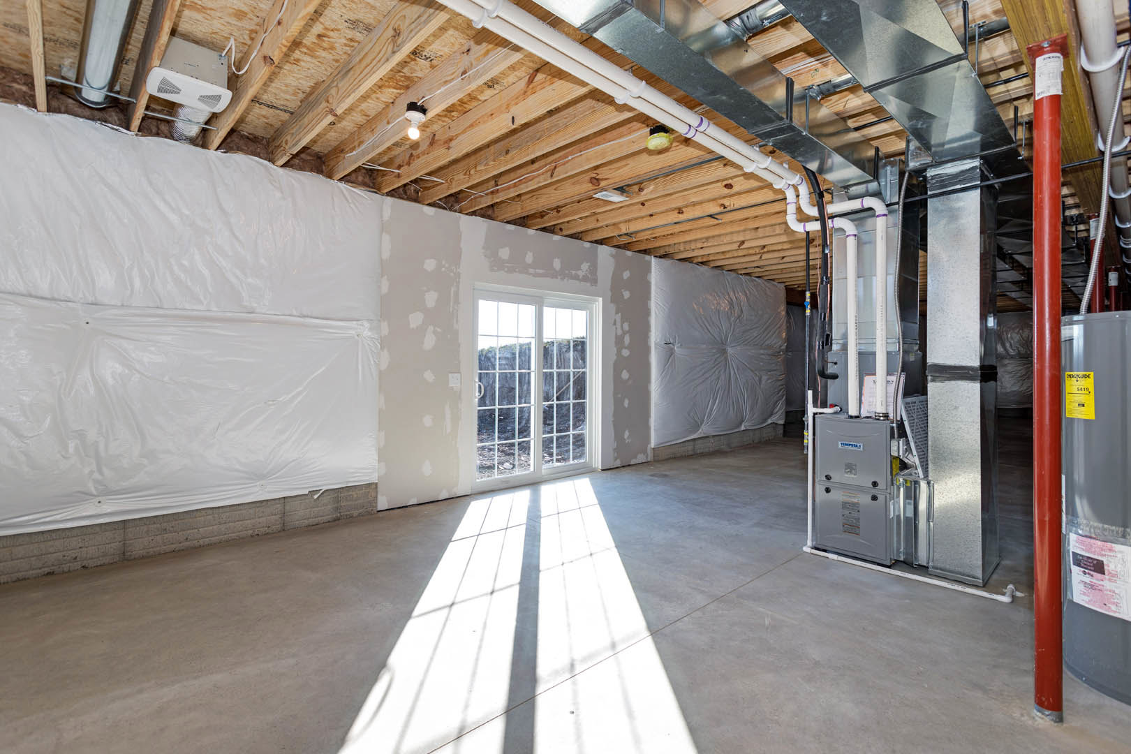 Spacious room featuring a large white wall, window with aluminum frame, light streaming in, exposed wooden beam, white plastic covering on wall, fire extinguisher near door