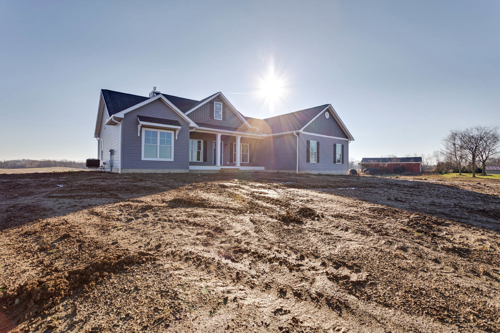 Modern house with white columns and large windows, set on a dirt hill with scattered trees and sunlight streaming through the partially finished roof