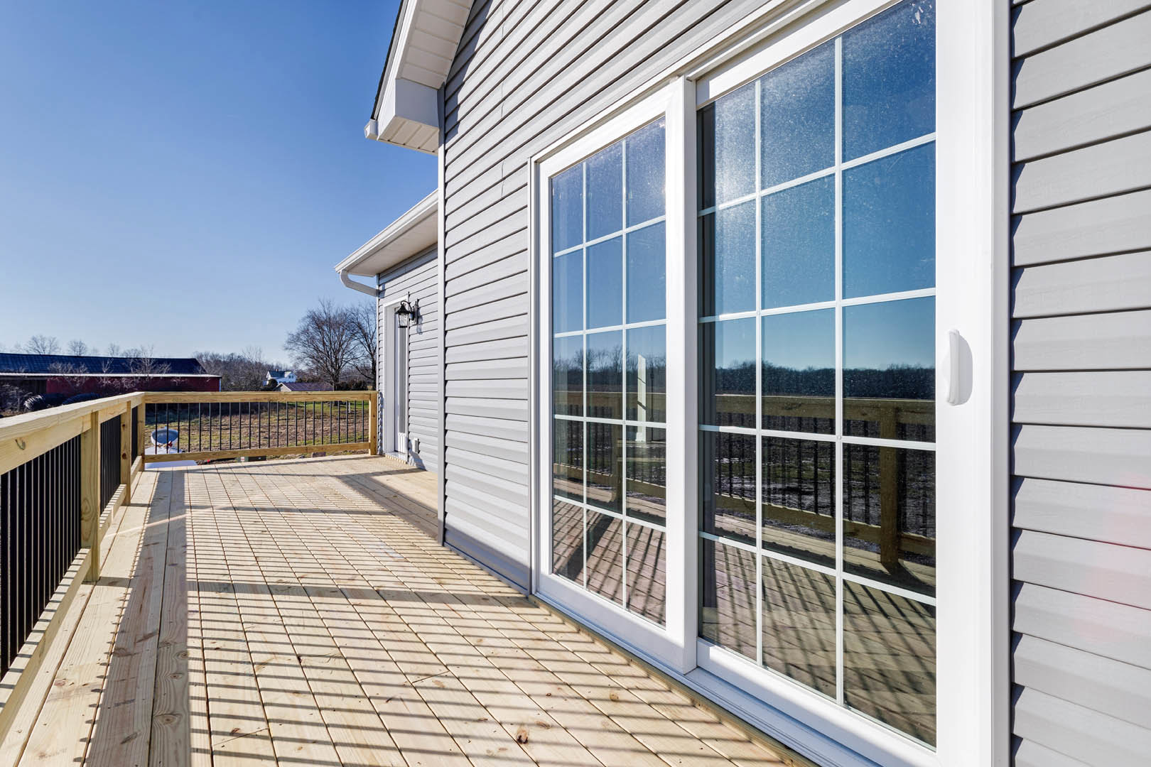 Wooden deck with metal railing, adjacent patio, large sliding glass door, blue roof, leafless tree, and expansive windows.