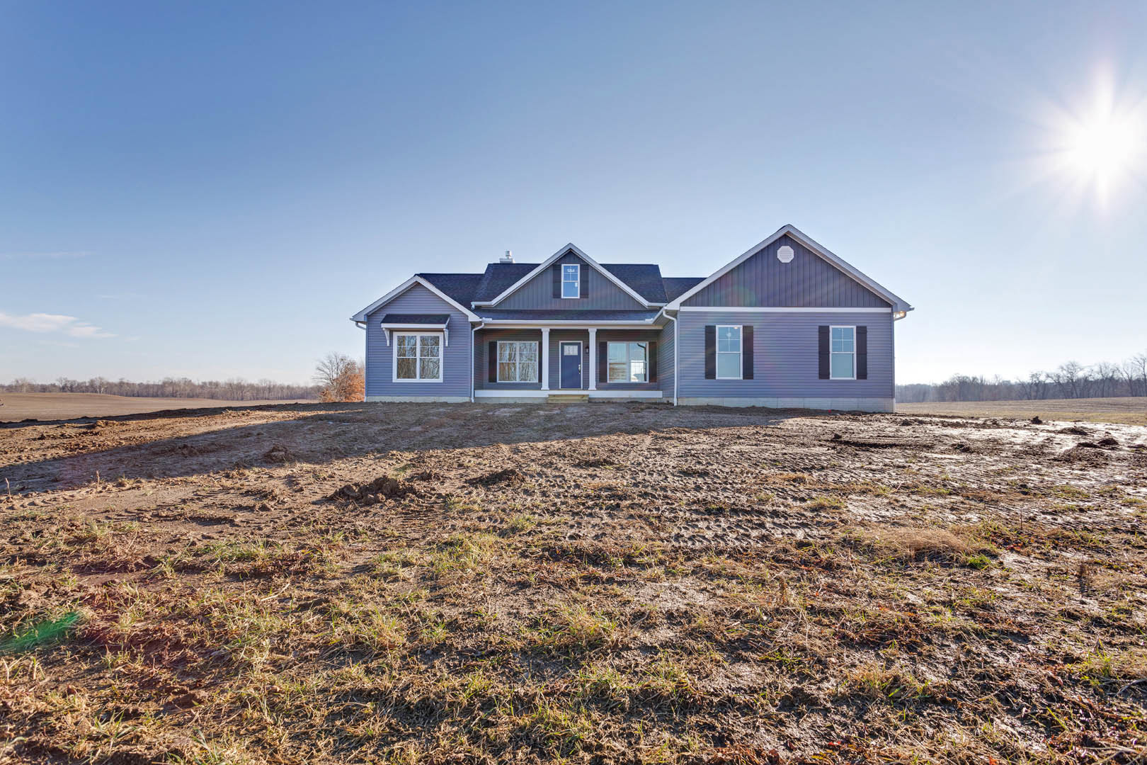 Two-story house with blue door and white-framed windows, gray roof, and large dirt field in front under clear blue sky