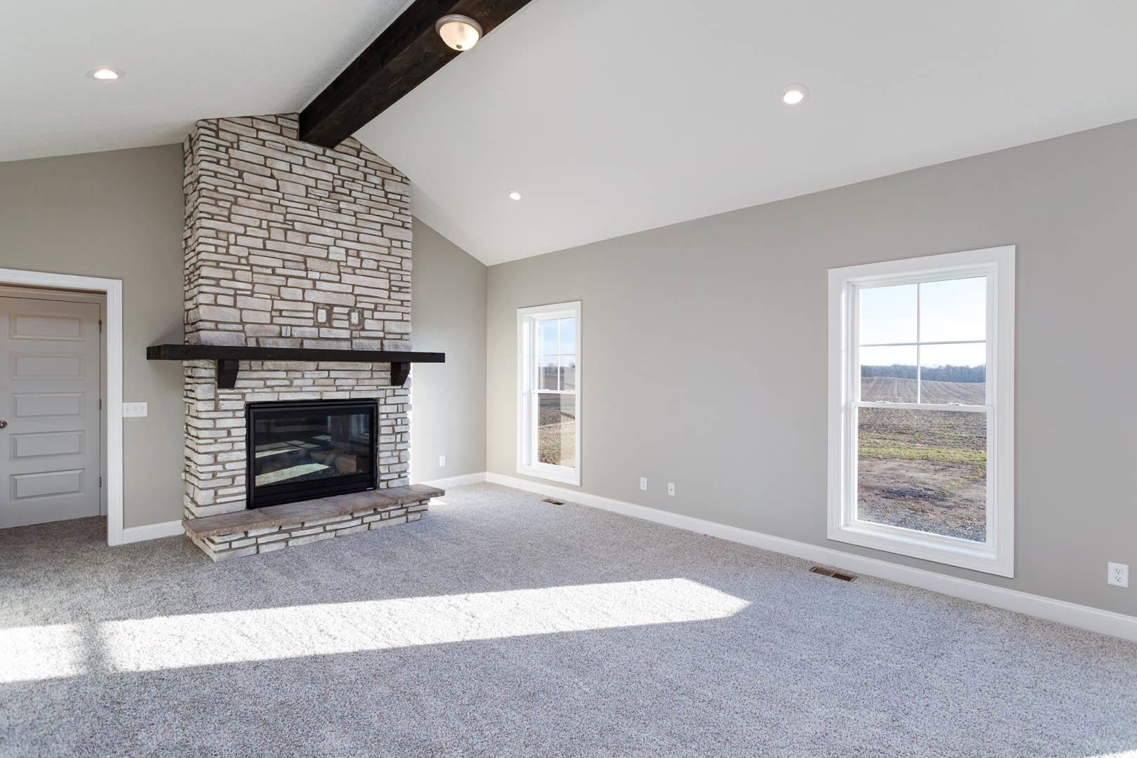 Carpeted living room featuring a stone fireplace, large windows overlooking a field, glass door reflecting stone wall, white walls, ceiling light fixture, and white interior door