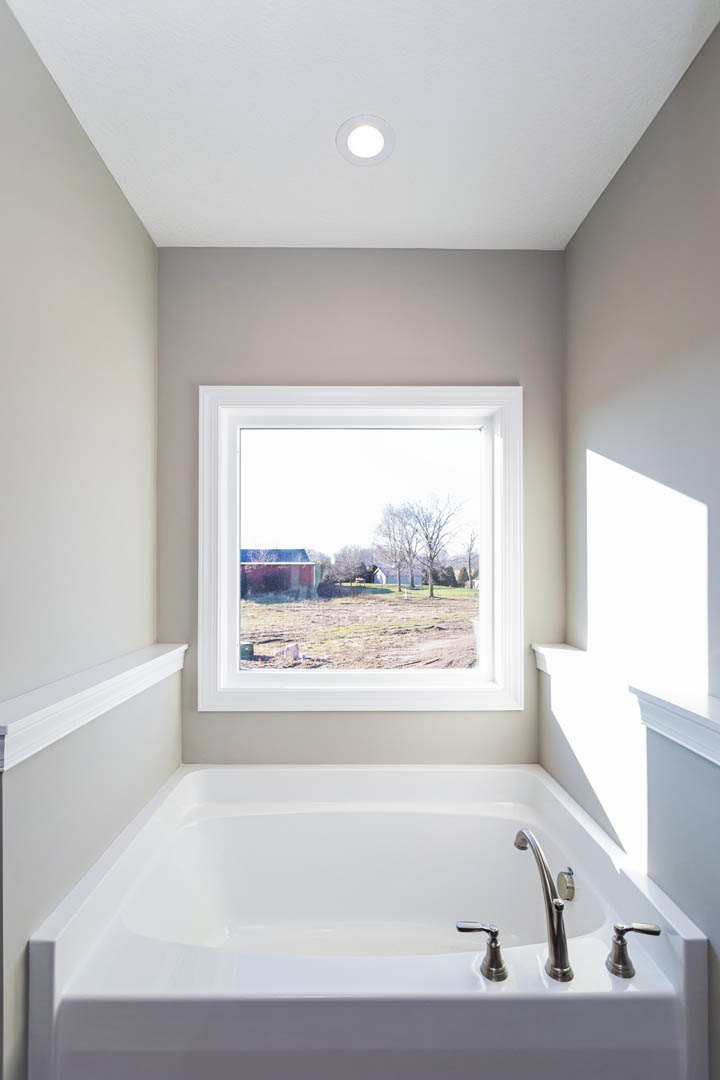 Freestanding white bathtub beneath large window overlooking farm fields and trees, chrome faucet, recessed ceiling light, minimalist bathroom design