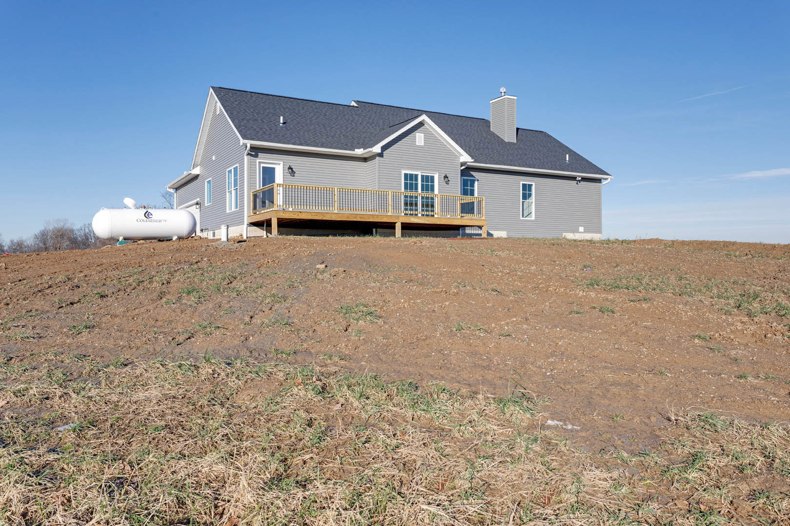 Two-story house with wooden deck and metal railing, white gas tank on grassy hillside, surrounded by rural landscape under blue sky