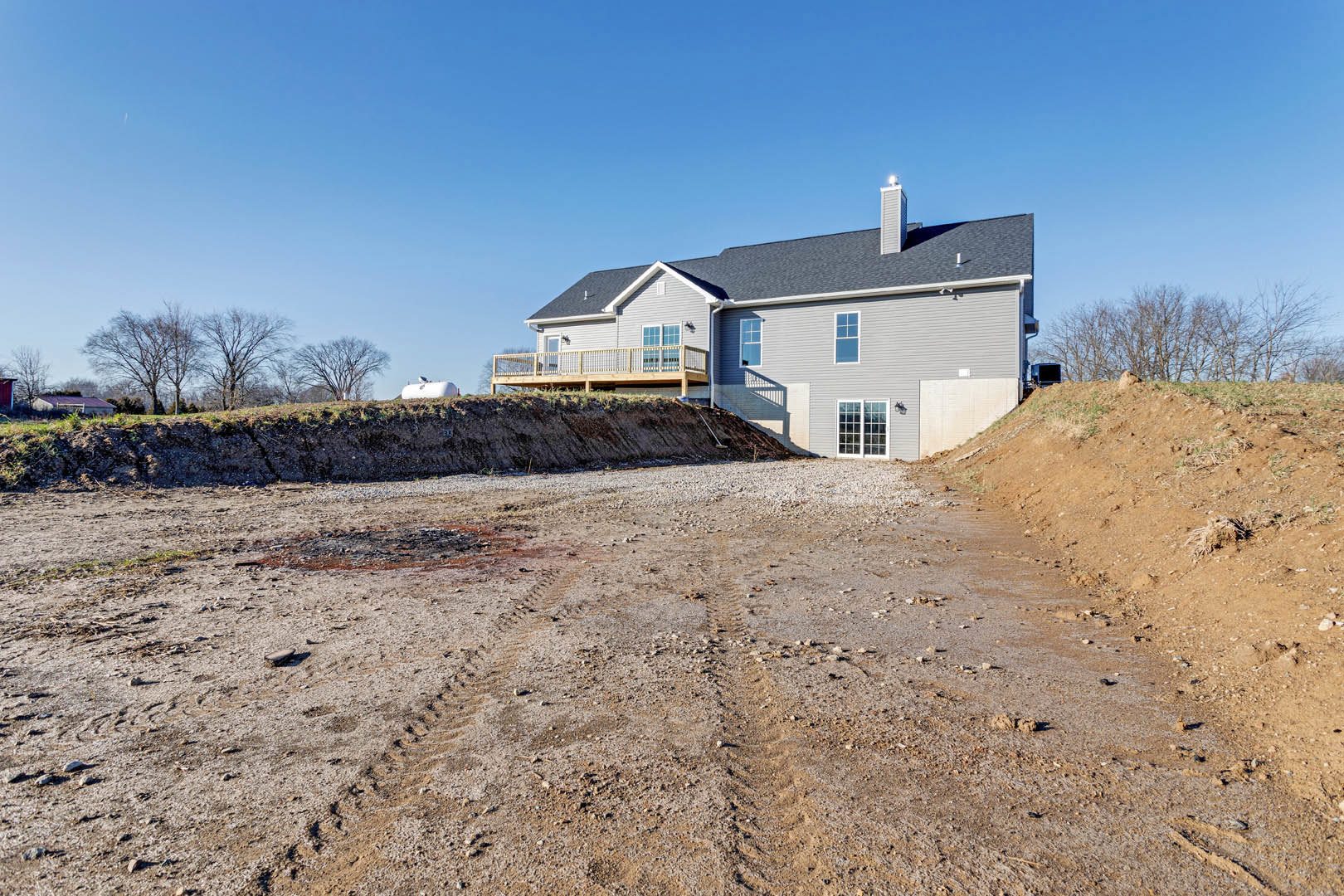 Two-story house with white double doors featuring glass panes, side wooden deck with light post, balcony above, tire tracks in dirt road leading to the home, grassy hill and