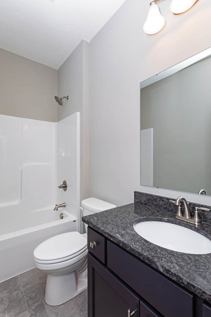 Bathroom with white tile walls, a white sink and faucet on a stone countertop, a toilet beside a built-in bathtub, and chrome plumbing fixtures.