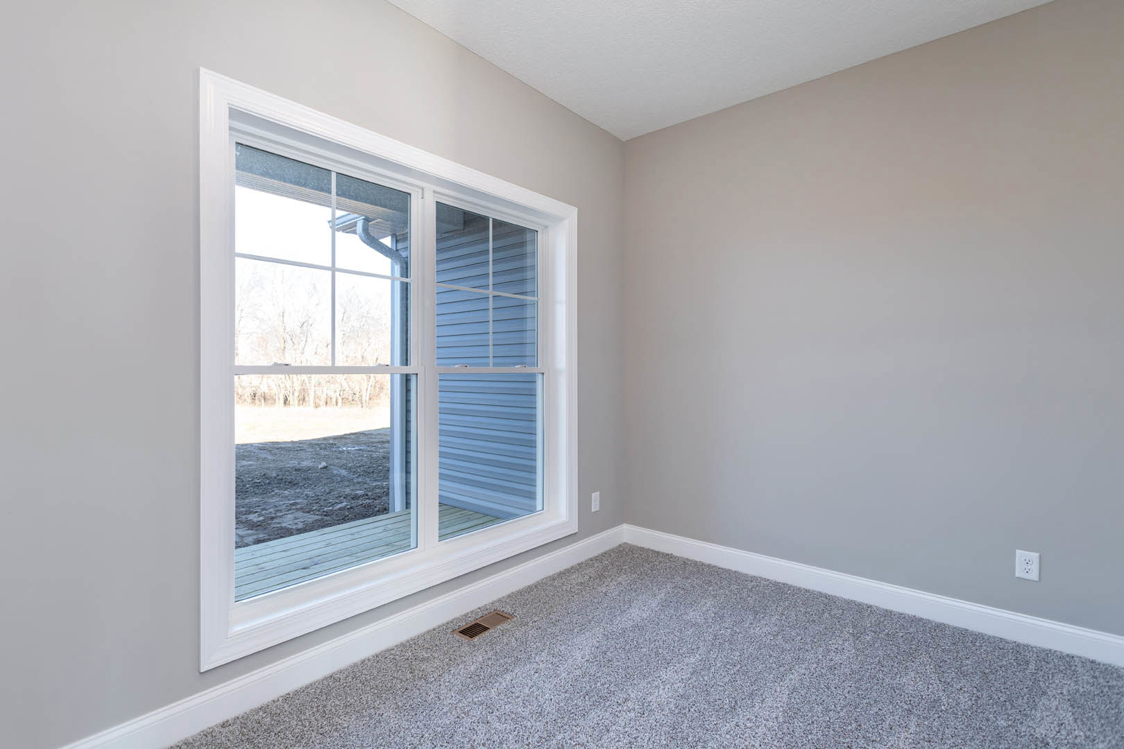 Bright room with white walls, large window overlooking grassy field, grey carpet flooring, wall vent near corner, simple window blind