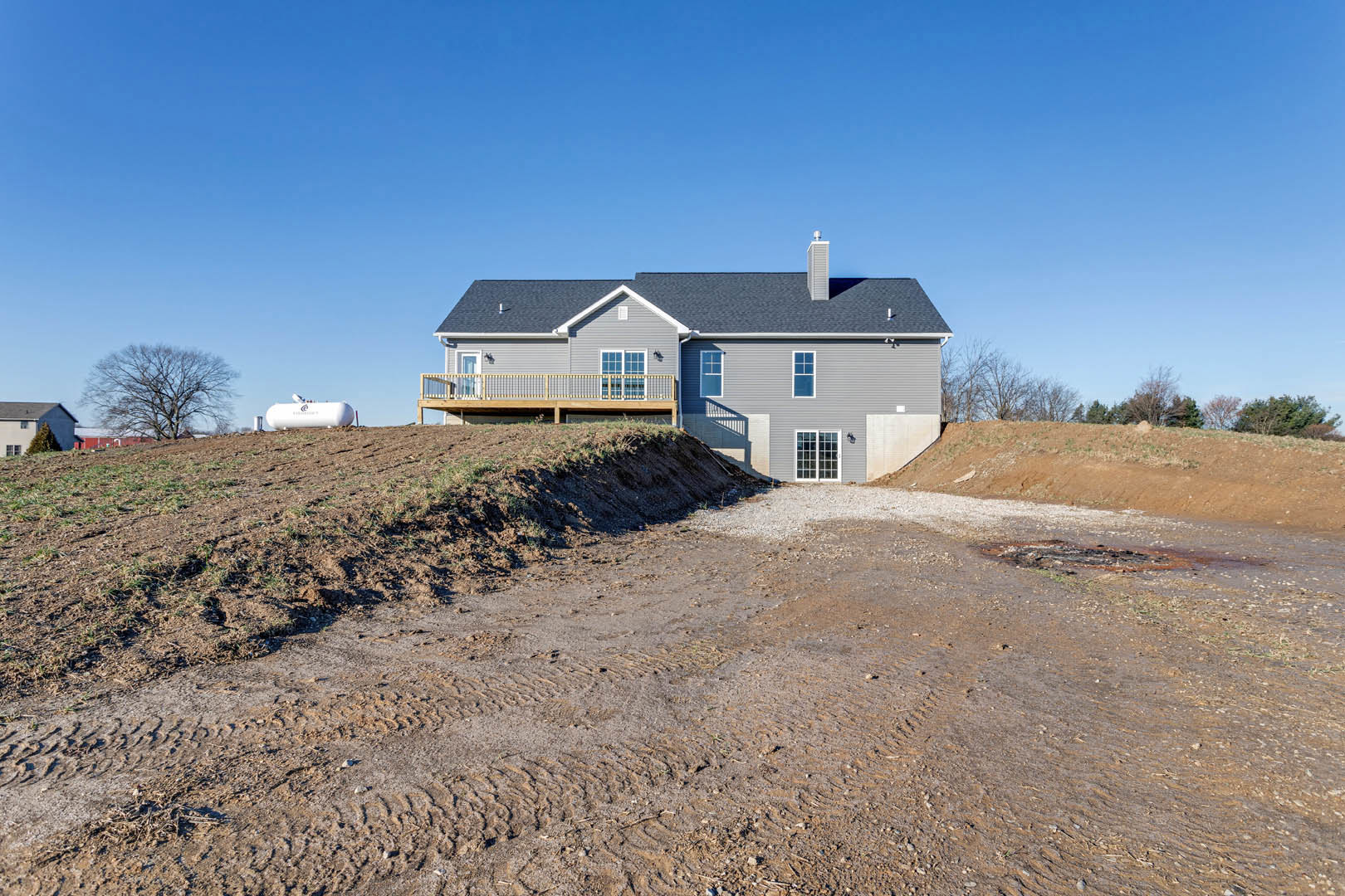 Two-story house with side deck and balcony, situated on a grassy hill next to a leafless tree, dirt road in foreground, cloudy sky above