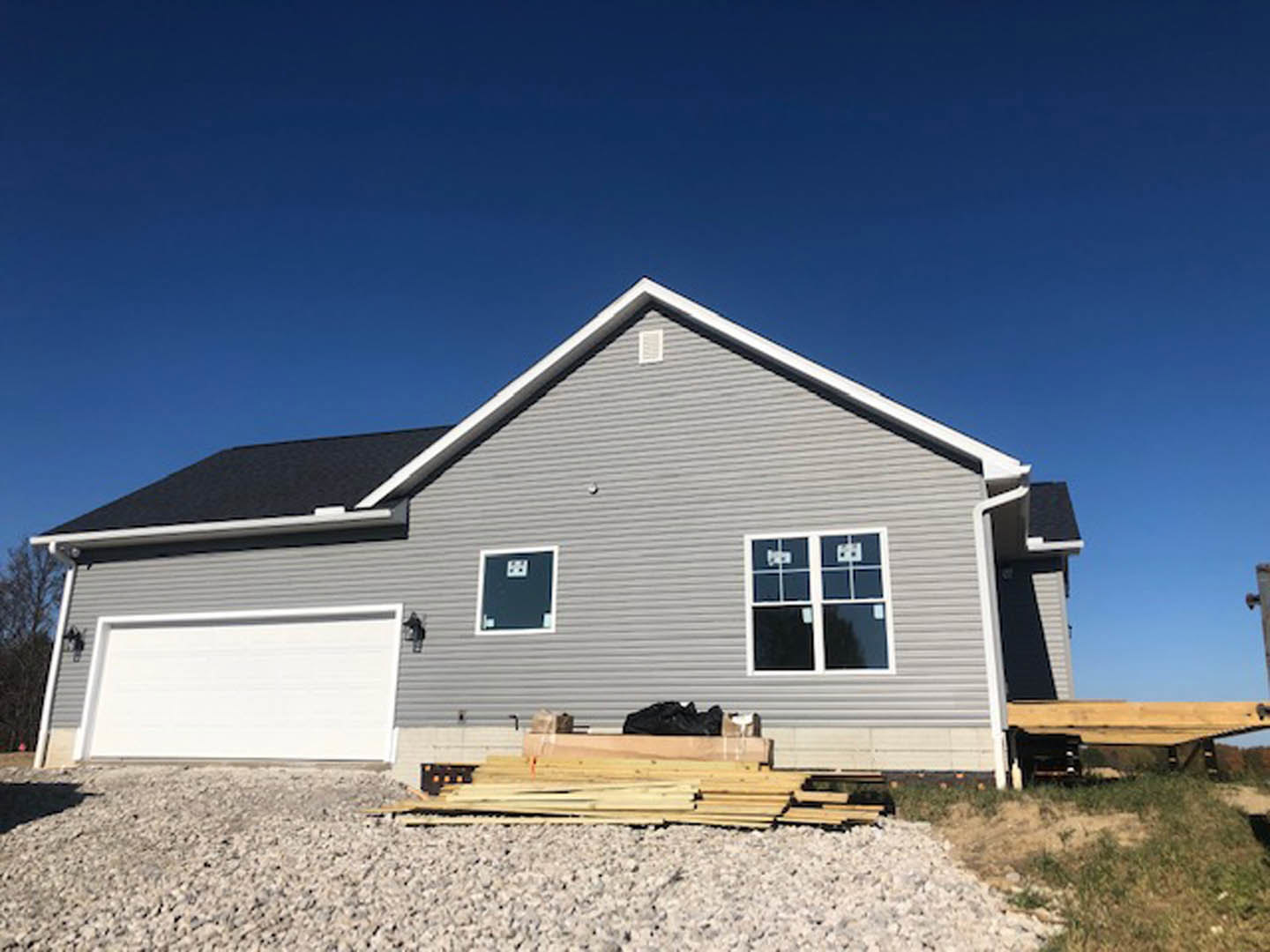 Framed house under construction with exposed wood beams, pile of lumber on gravel in foreground, historic Rockingham Meeting House visible in background, open field and blue sky