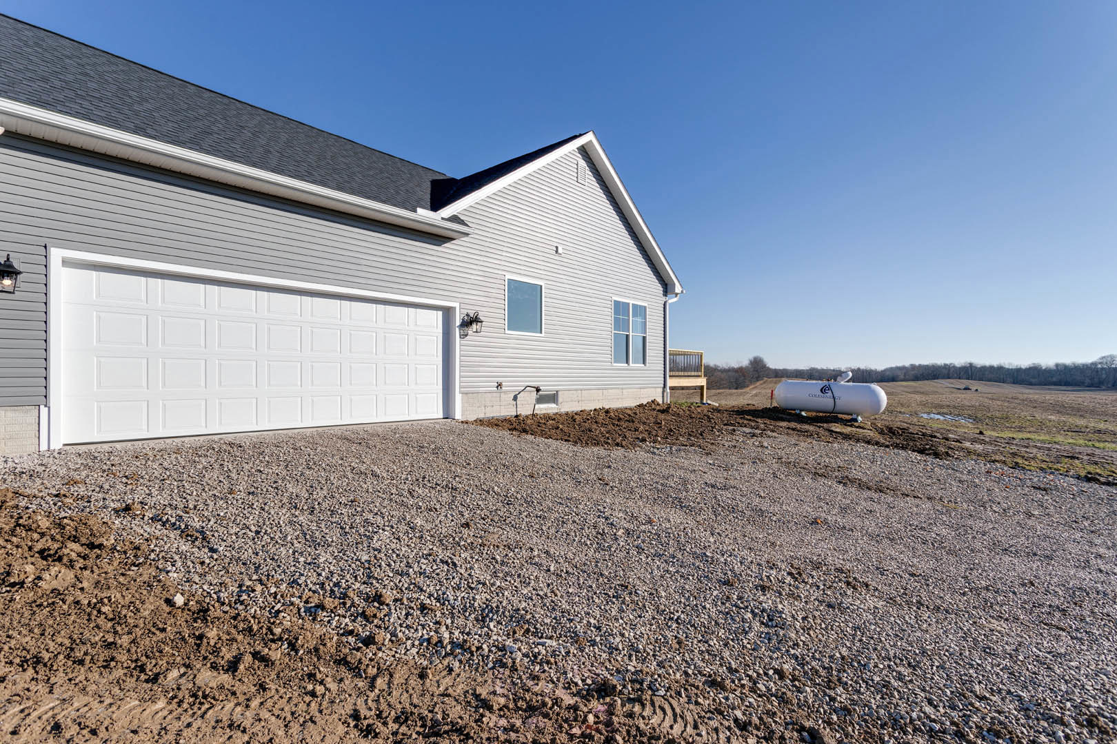 Gravel driveway leading to a white garage with grey roof, large cylindrical white tank beside siding-clad house, single window visible on building exterior