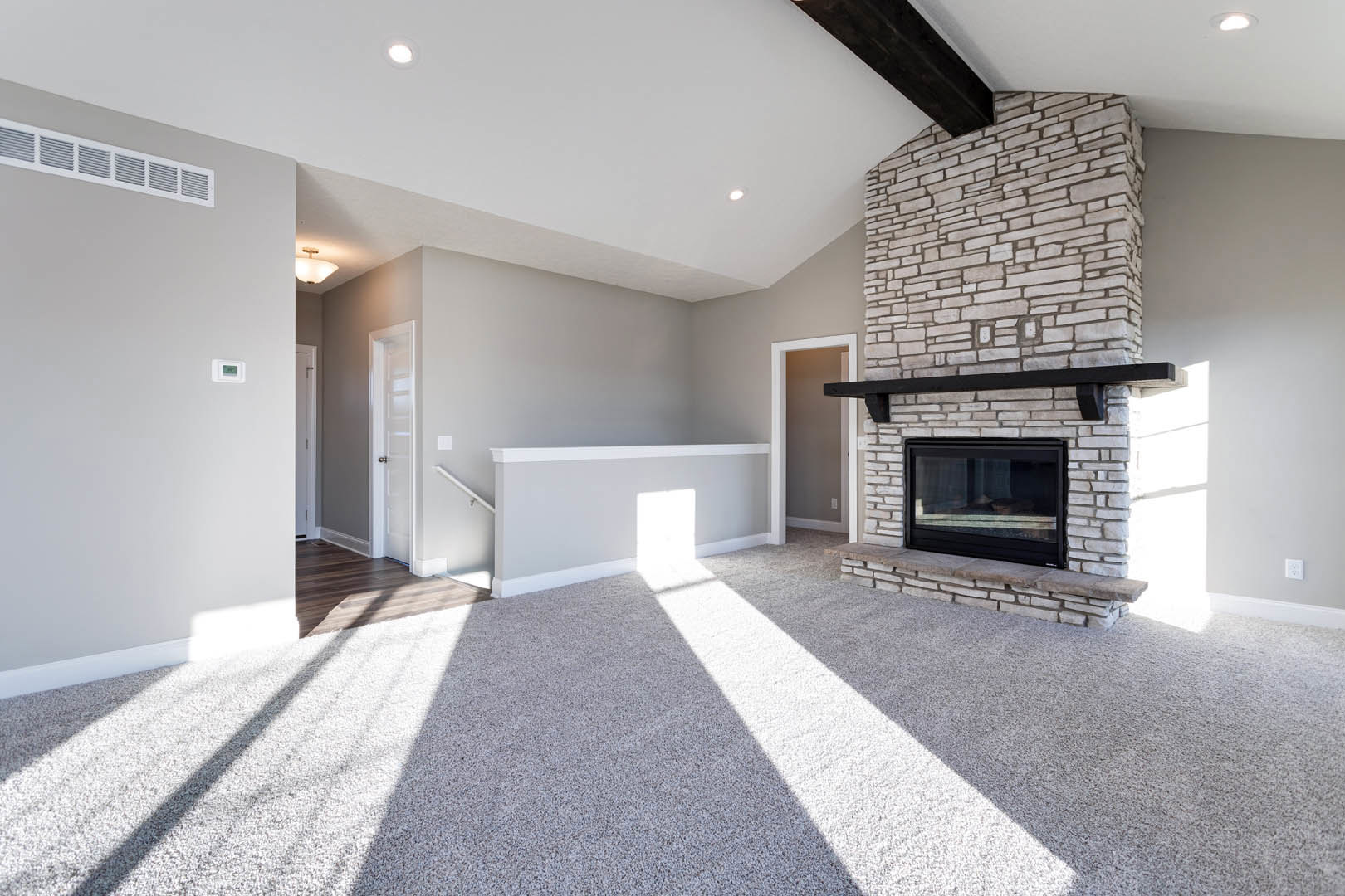 Living room with beige carpet, stone fireplace with glass door, white walls, ceiling light fixture, sunlight streaming through window, white door leading to adjacent space, black