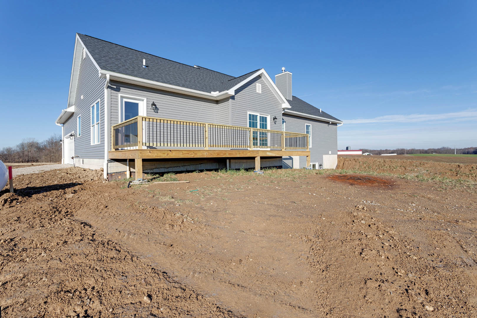 Two-story house with large windows, wooden deck featuring metal railing, fenced yard bordered by a dirt field, cloudy sky overhead