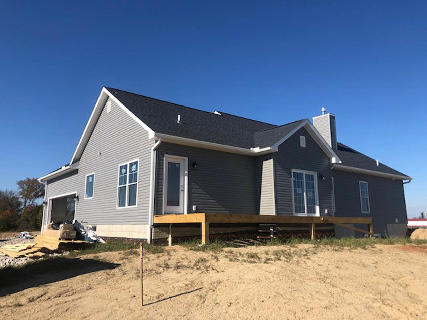 Two-story house under construction with exposed siding, covered porch, dirt yard, wooden bench on grass, and visible windows and doors