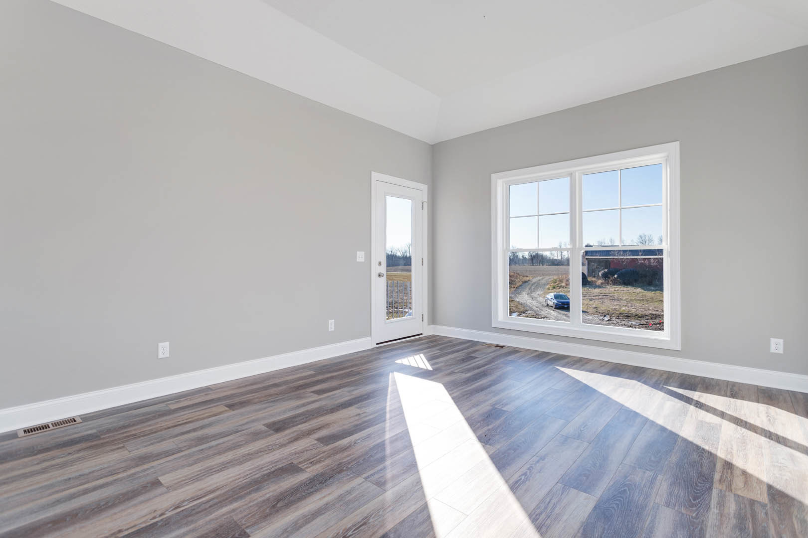 Sunlit wood flooring in a room with white plaster walls, large window, and white door opening to an outdoor view.