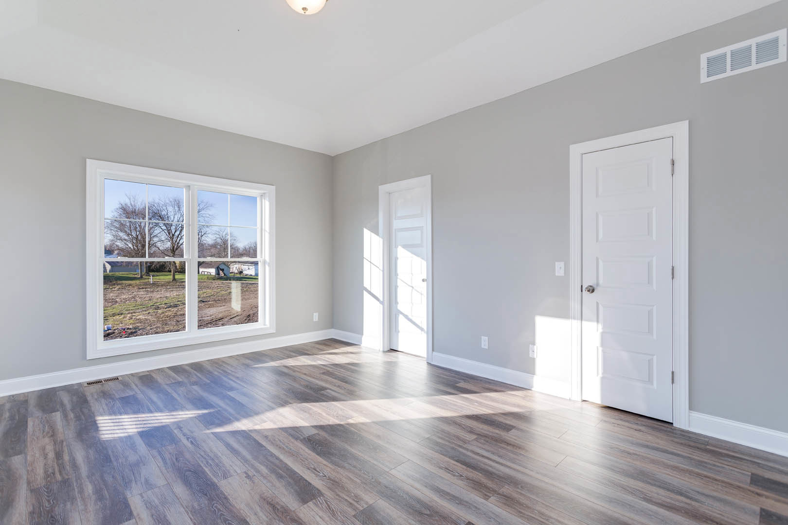 Sunlit room with wood flooring, white walls, a white door with silver knob, and a window overlooking a field and trees