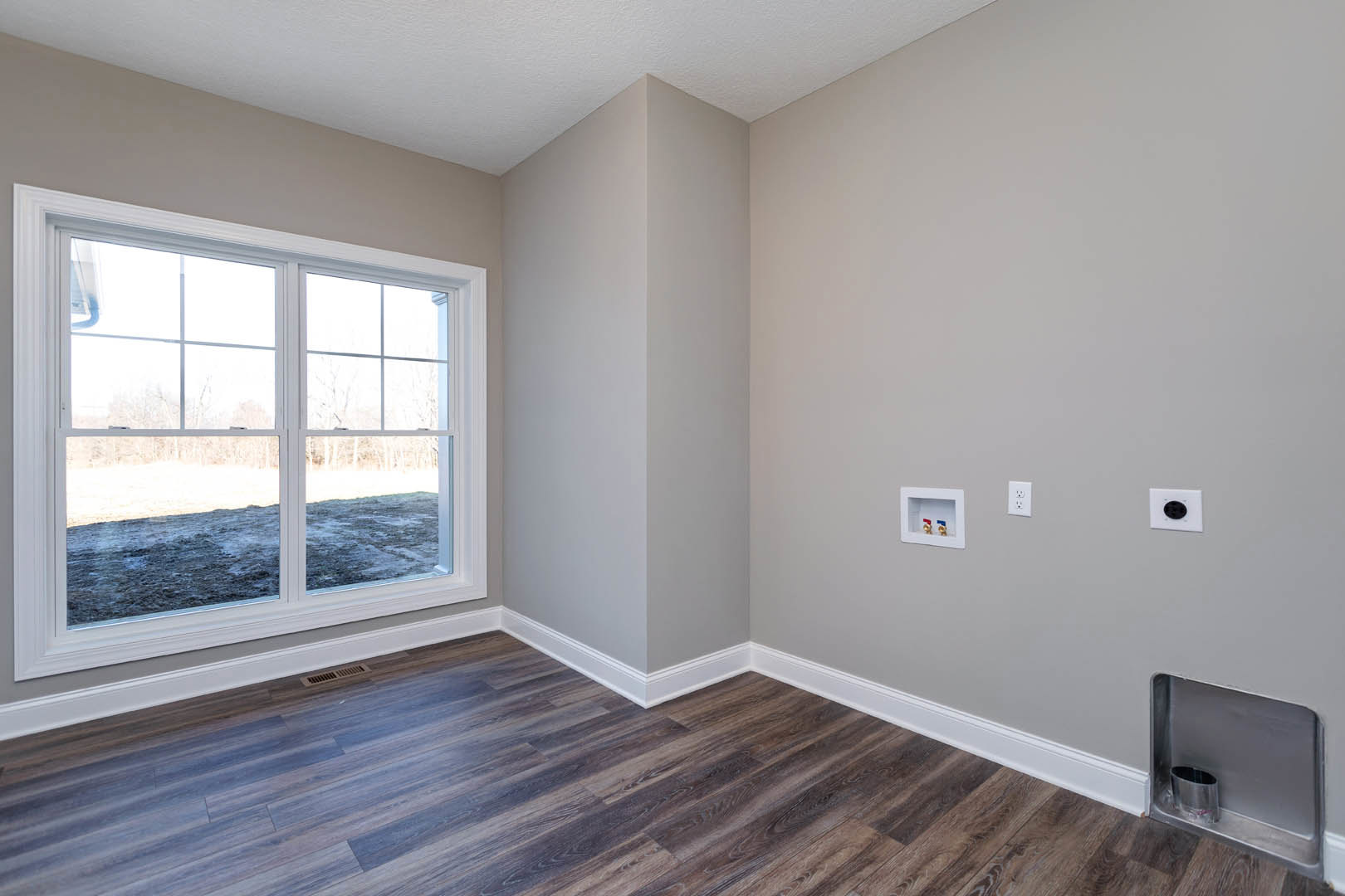 Sunlit room with large window, light wood flooring, white plaster walls, visible wall outlet, and a recessed metal container with valves.