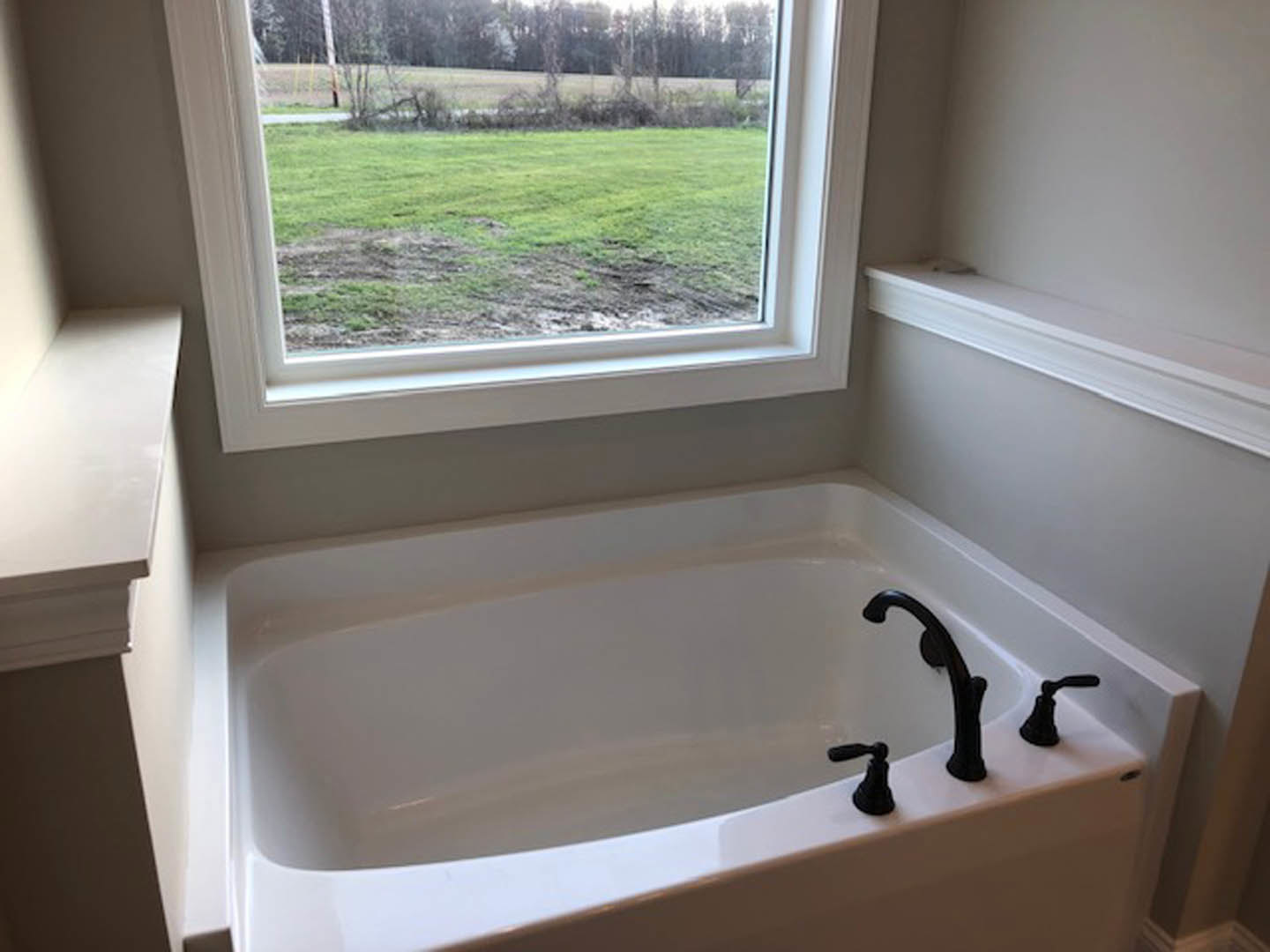 Freestanding bathtub with black faucet beneath large window overlooking green grass
