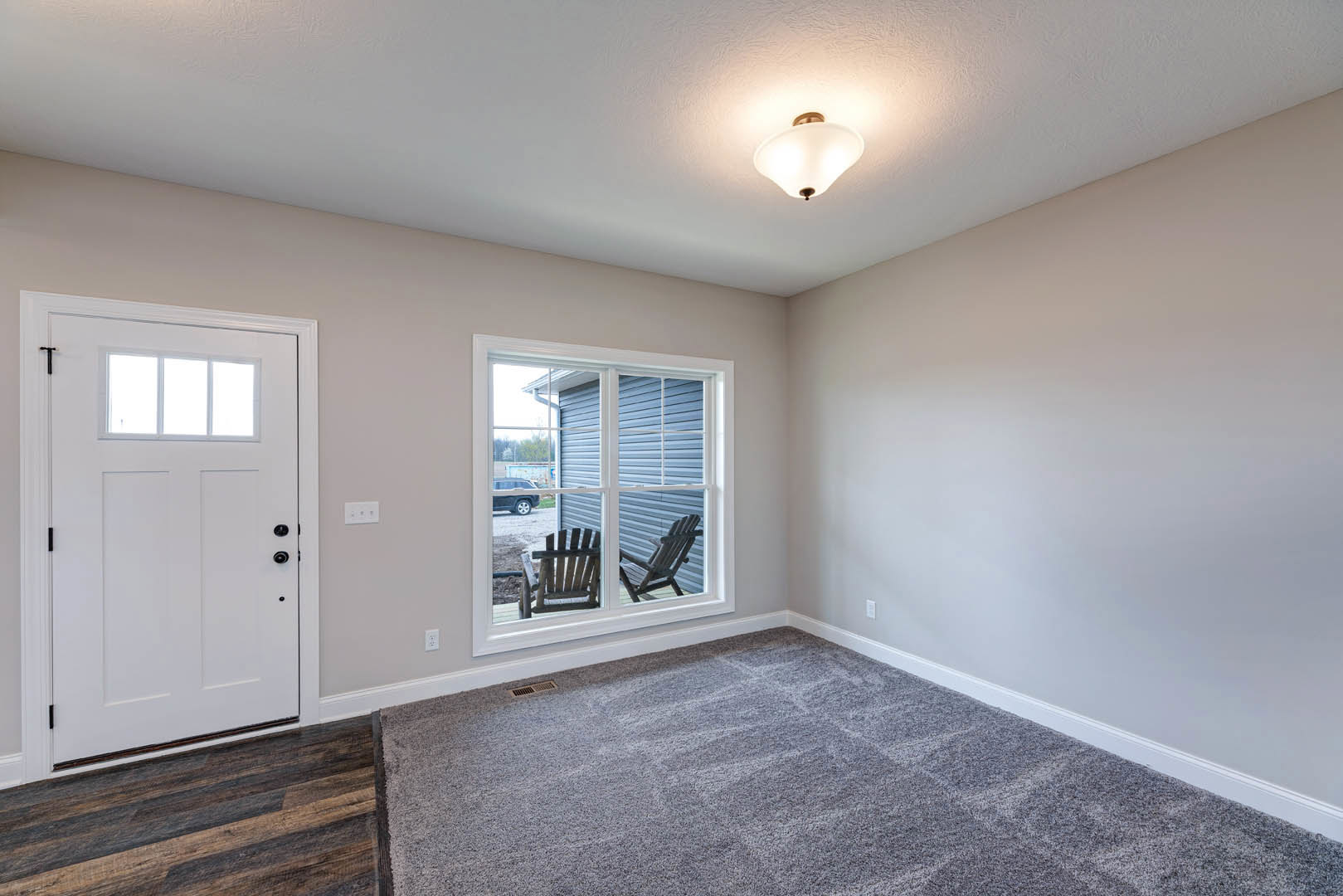 White paneled door beside a large window, grey carpeted floor, two upholstered chairs, white plaster walls, ceiling light fixture, wood flooring visible in foreground