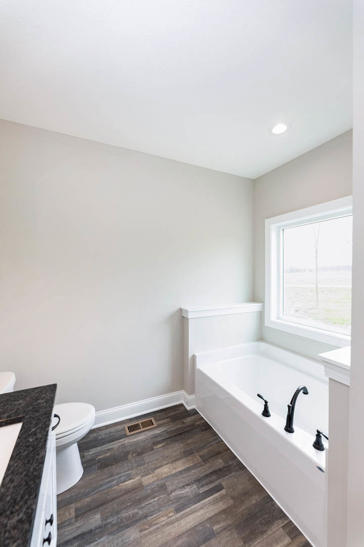 Bathroom with freestanding bathtub, white toilet, rectangular sink on black countertop, wood flooring with floor vent, large window overlooking grassy field, light tile walls